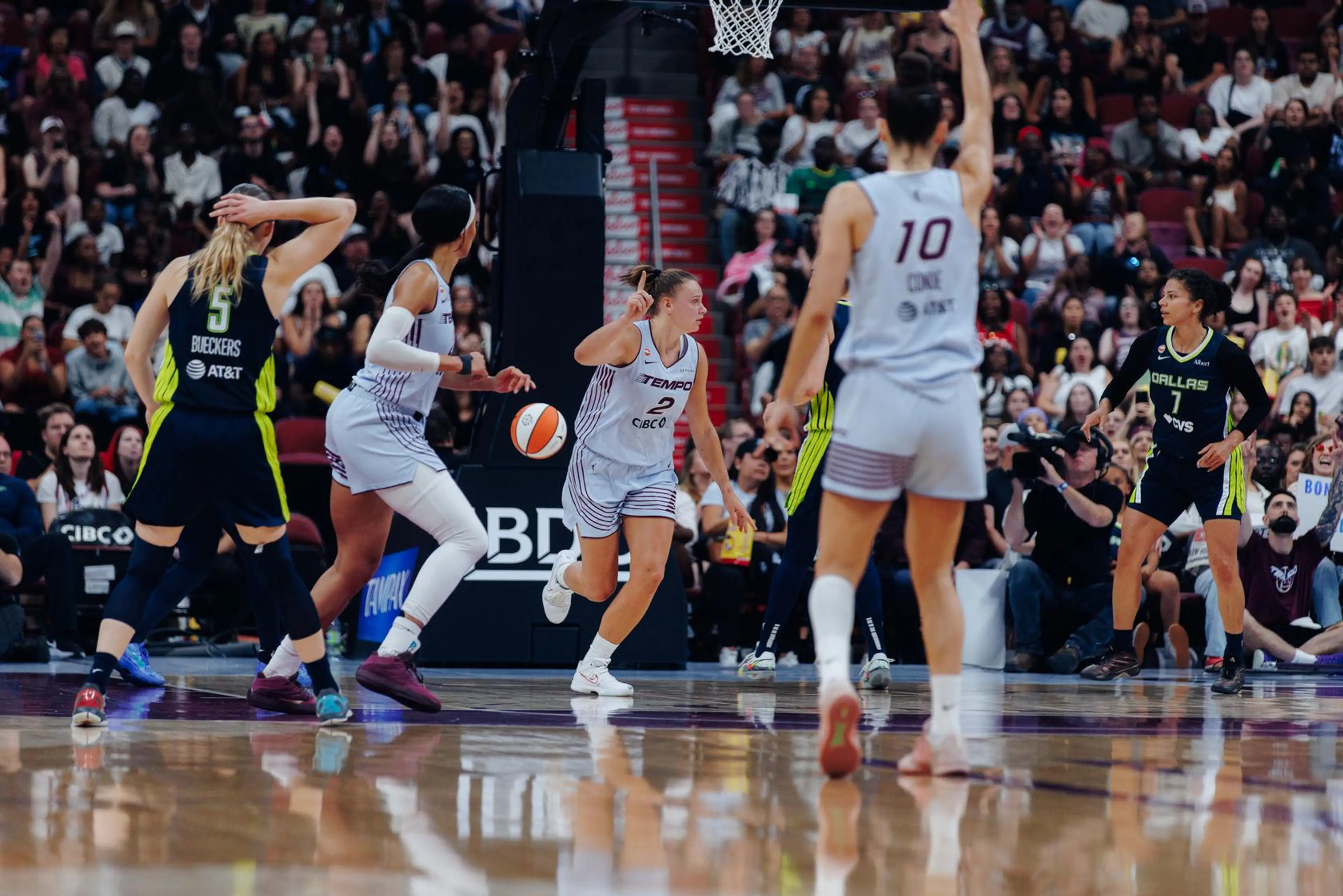 Five basketball players on court during a game, three in white uniforms and two in black uniforms, with a crowd in the background.