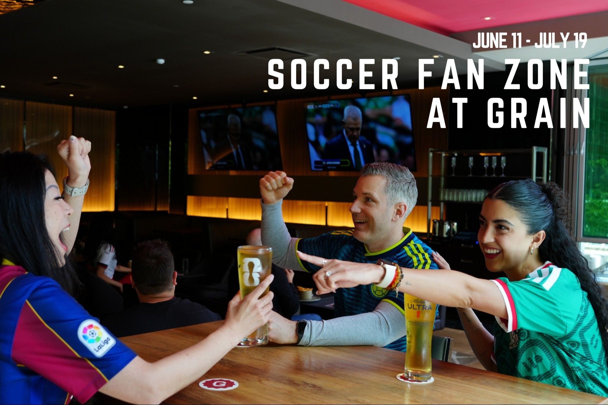 Three people at a bar cheering and holding glasses of beer, watching a soccer game on TV screens in the background.