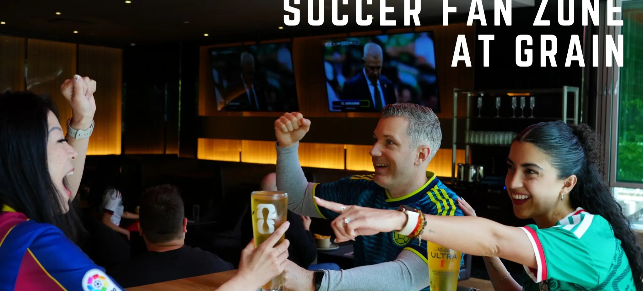 Three people at a bar cheering and holding glasses of beer, watching a soccer game on TV screens in the background.