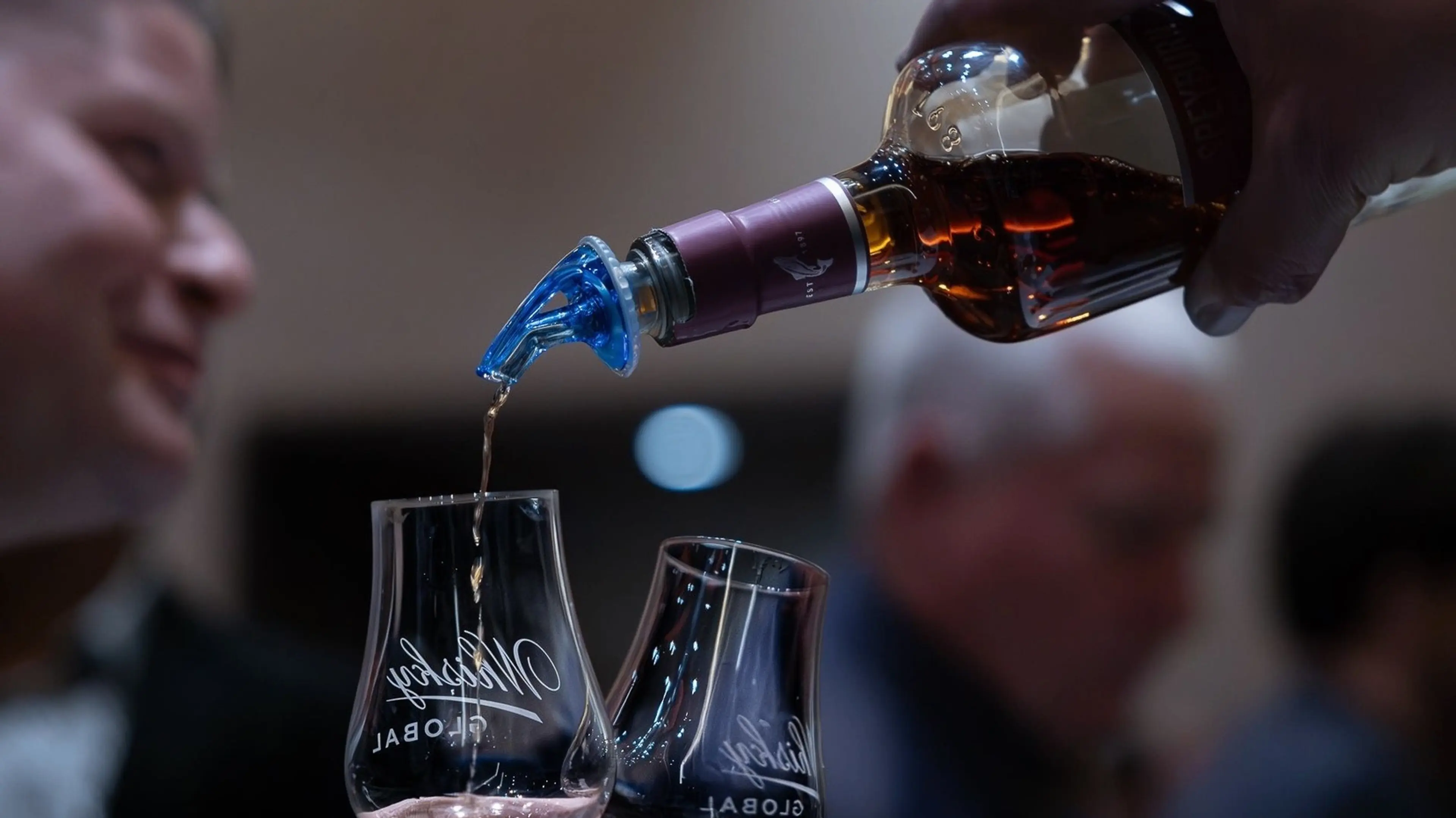 A person pouring whiskey from a bottle into a glass with another glass beside it, people blurred in the background.