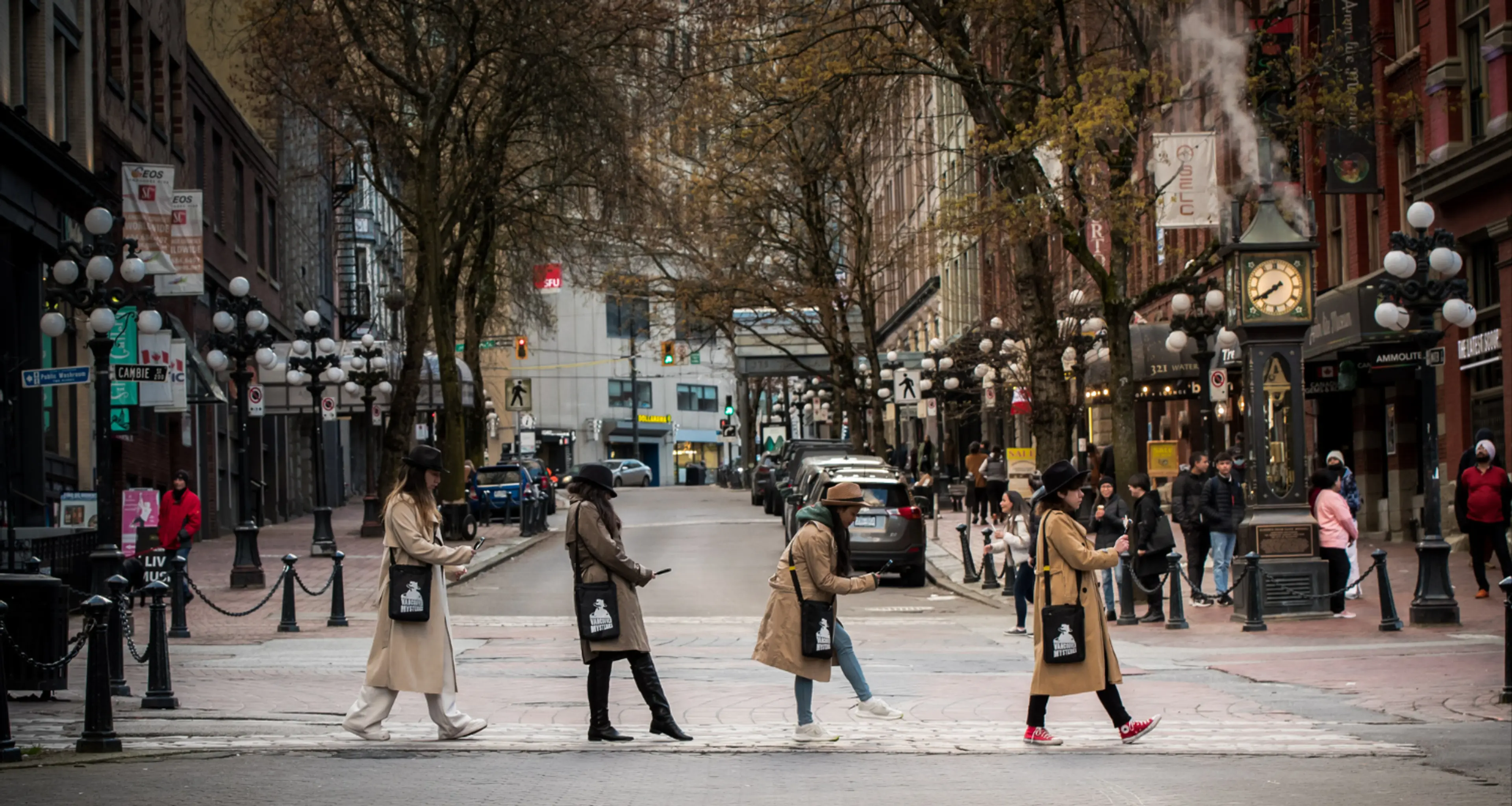 Four people in coats and hats crossing a street in a city with a steam clock and pedestrians in the background.