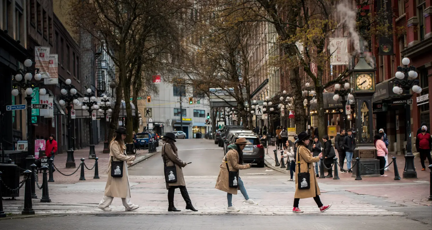 Four people in coats and hats crossing a street in a city with a steam clock and pedestrians in the background.