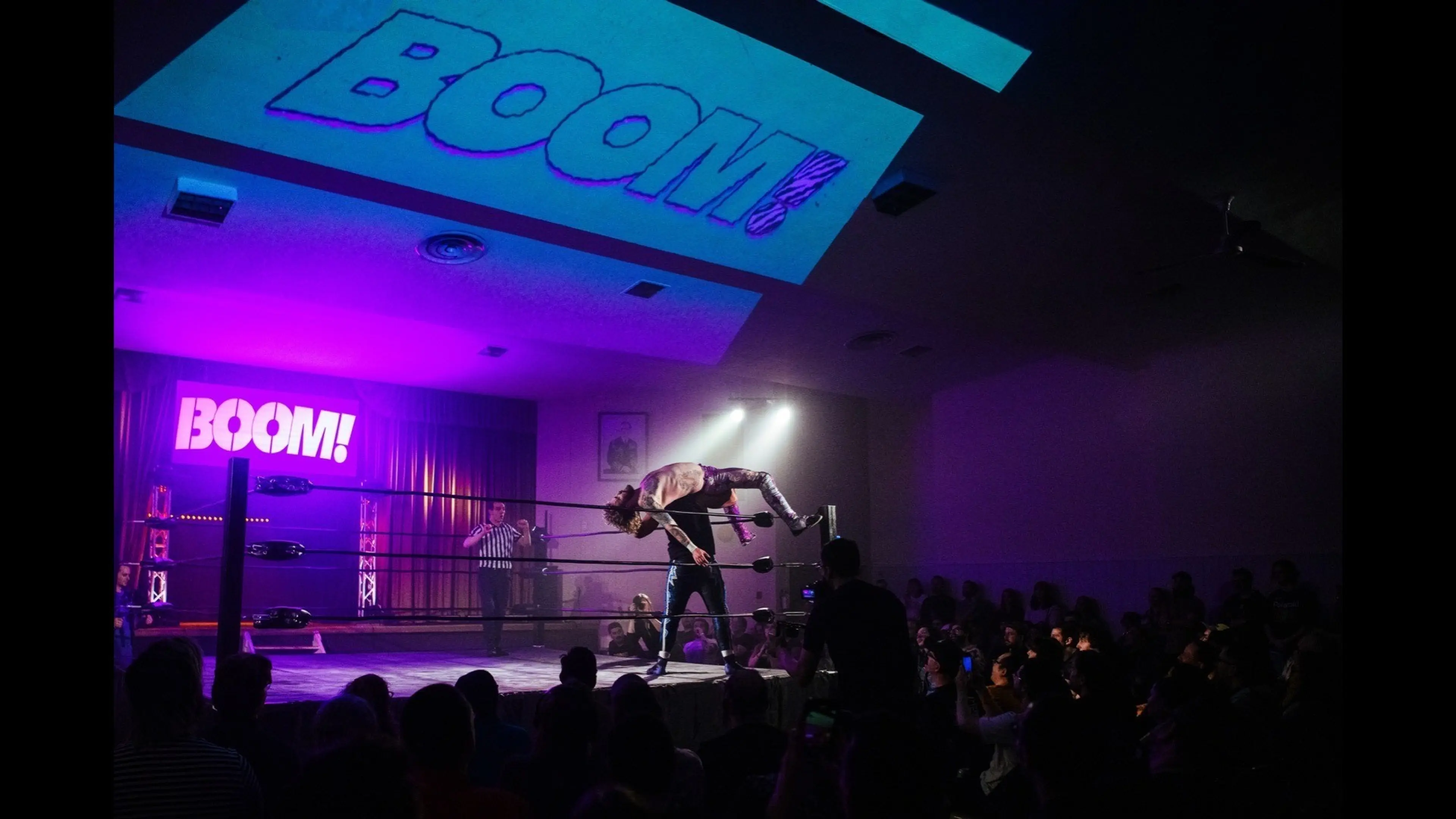 Wrestling match with one wrestler lifting another inside a ring, audience watching, and the word BOOM! lit up in purple on the wall.