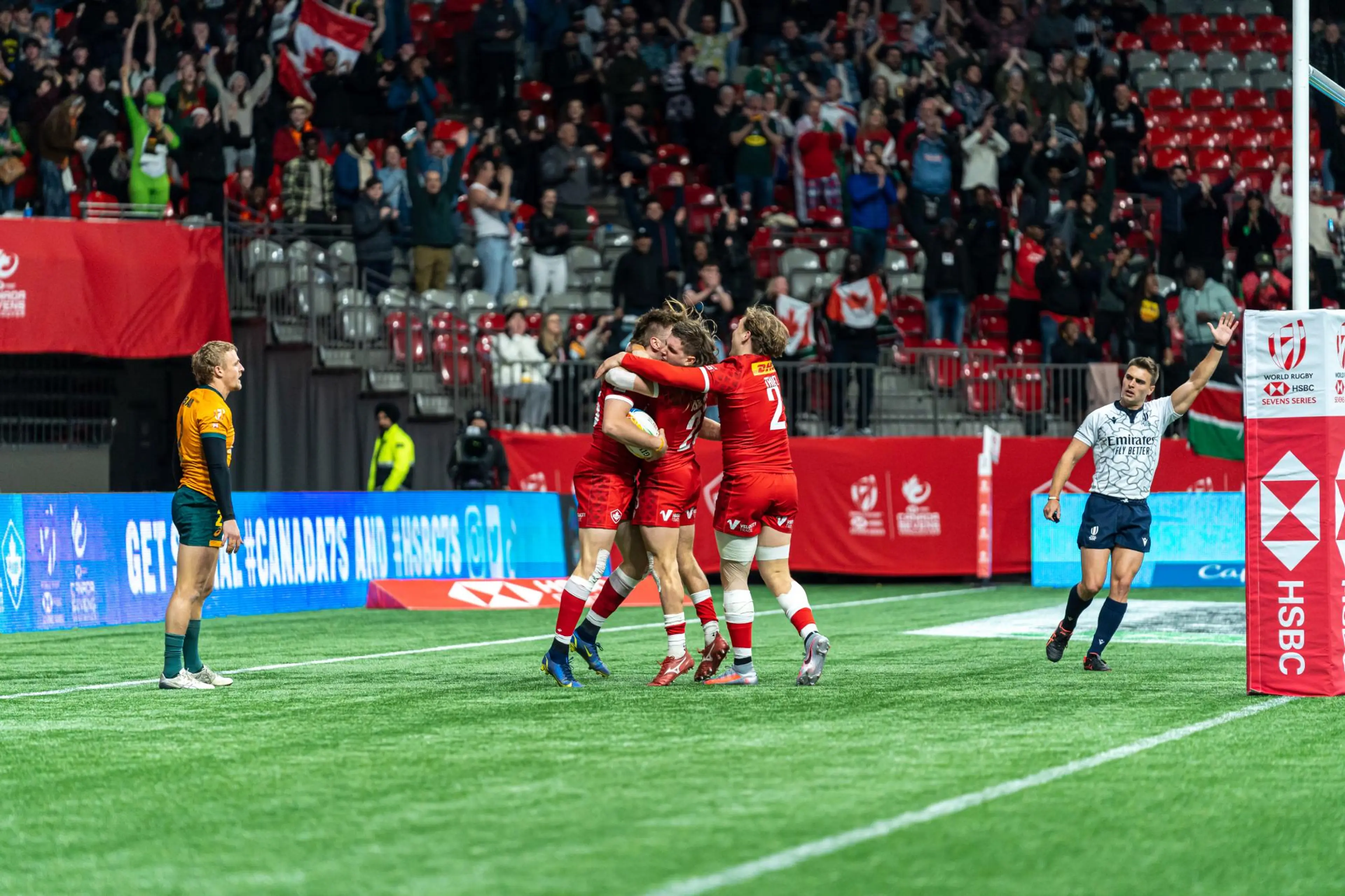 Rugby players in red celebrating a try near the goalpost while an opponent in orange and a referee in white watch.