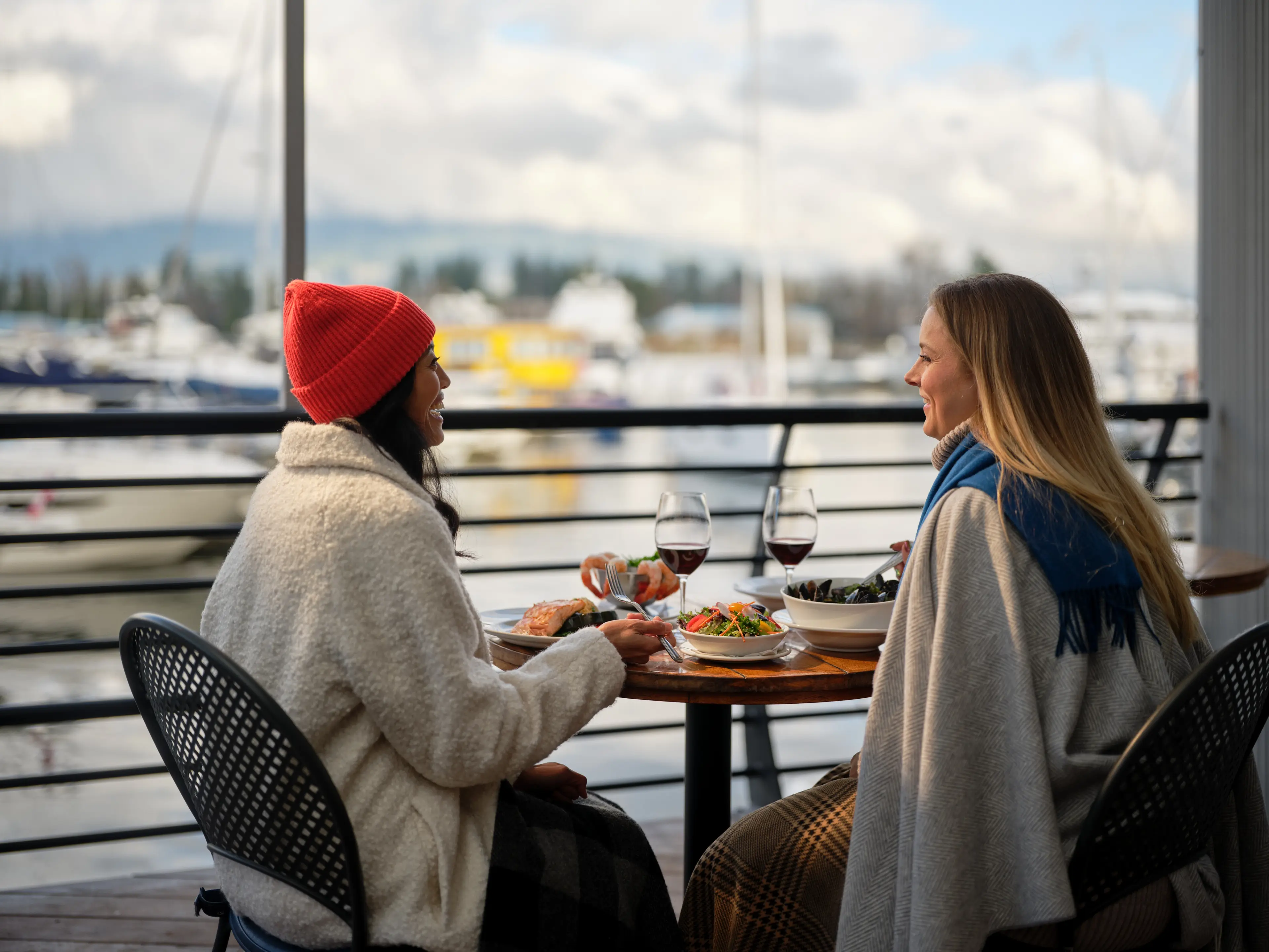 Two people sitting at a table outdoors near a marina, enjoying a meal with wine glasses, one wearing a red beanie and white jacket, the other with long hair and a blue scarf.