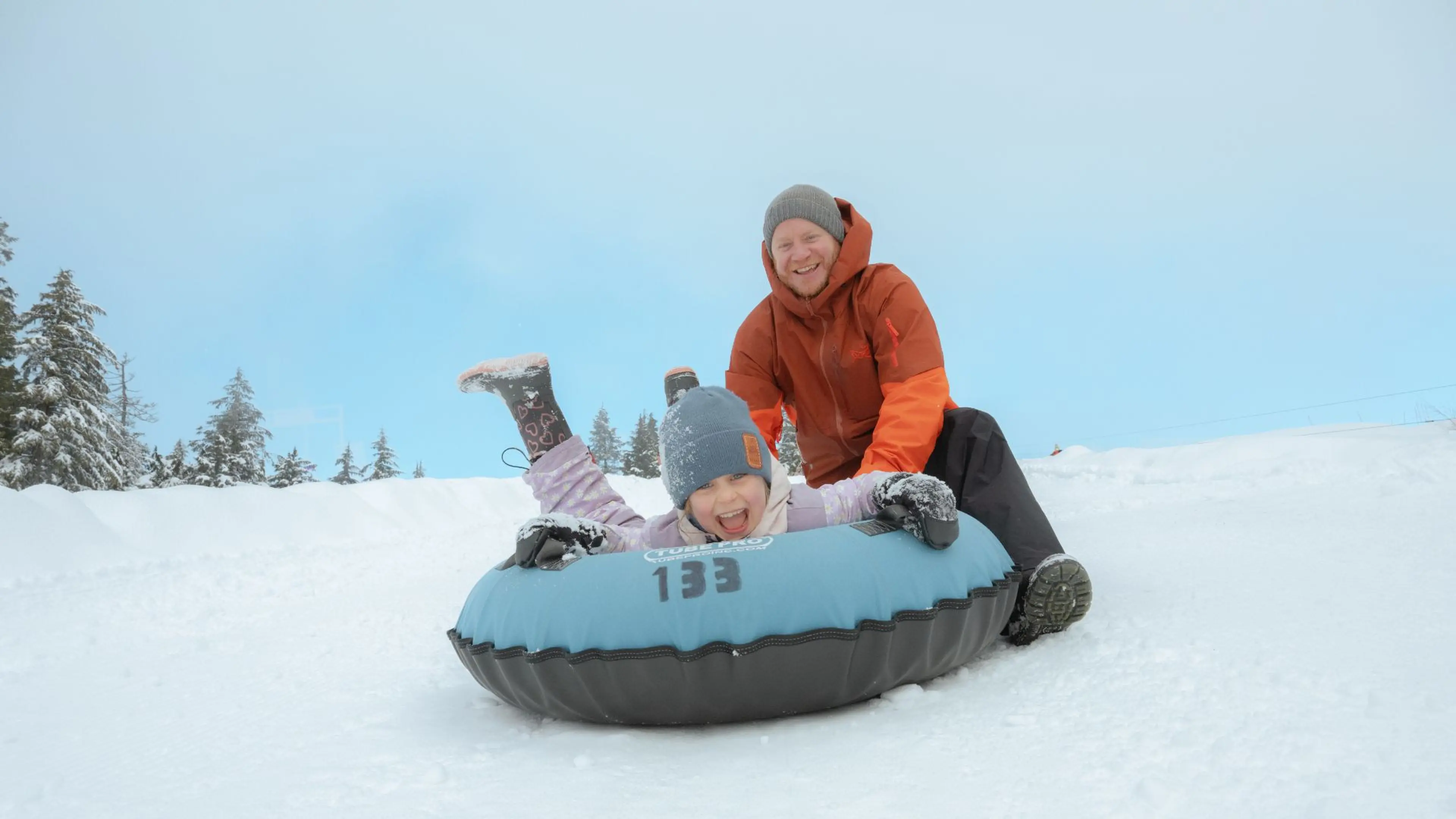 Child in a blue hat and winter clothes sliding on a blue snow tube with an adult in an orange jacket pushing from behind on a snowy hill.
