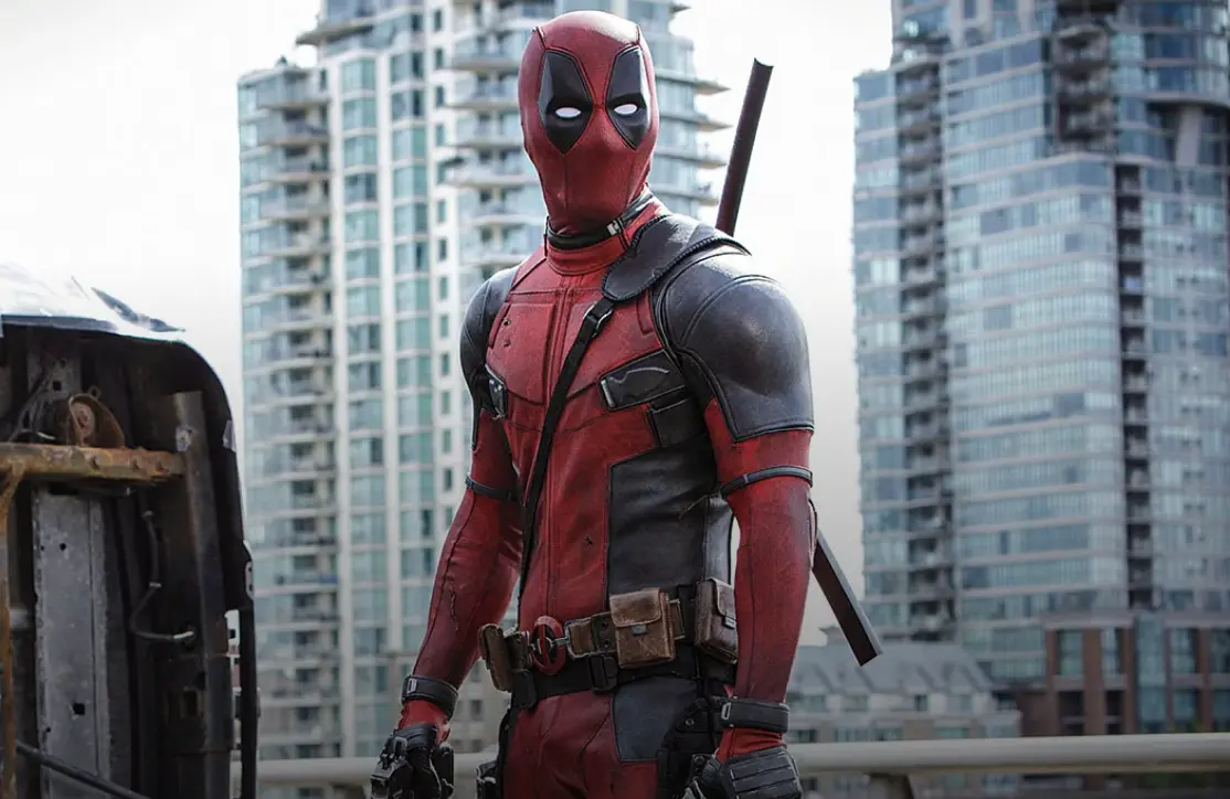 A person in a red and black superhero costume with swords strapped to their back stands next to an overturned vehicle on a city street in Vancouver, tall buildings rising behind them—perhaps searching for free wifi Vancouver offers nearby.