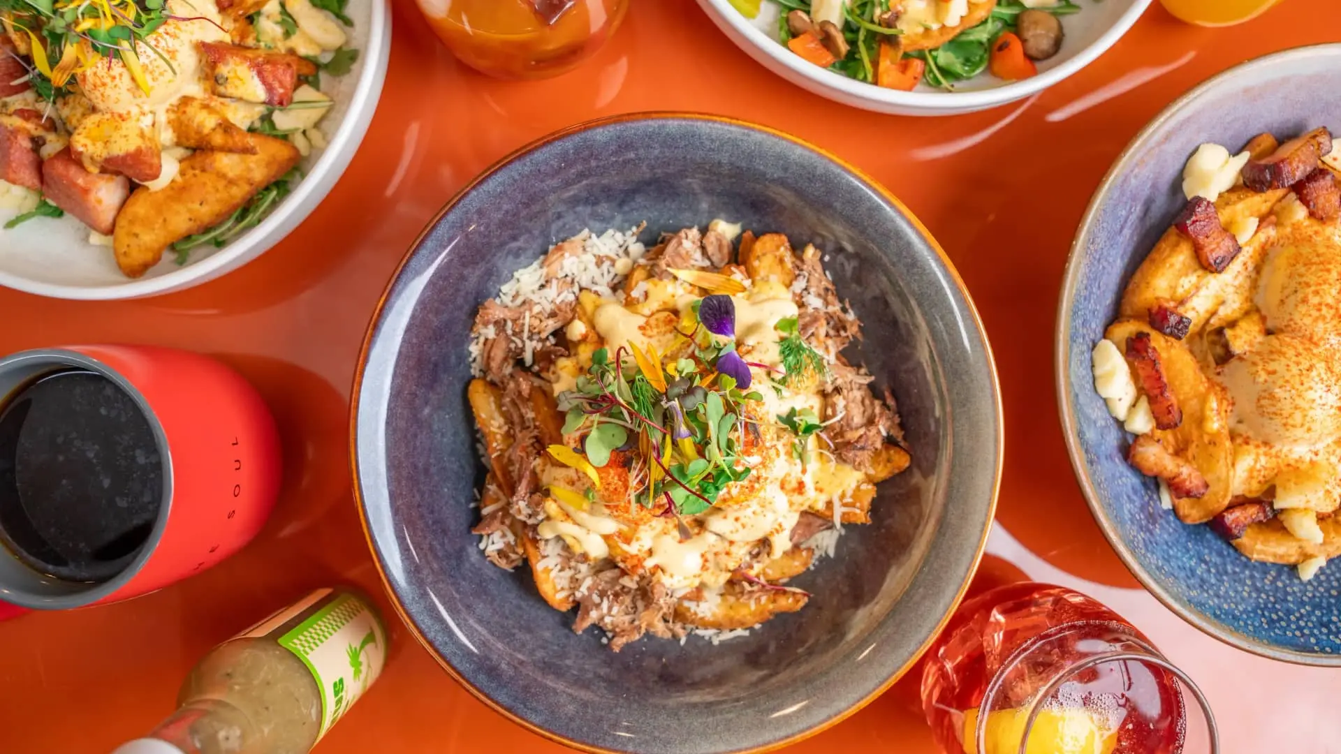 Bowls of loaded fries with cheese, meat, and greens on an orange table with drinks and a red cup.