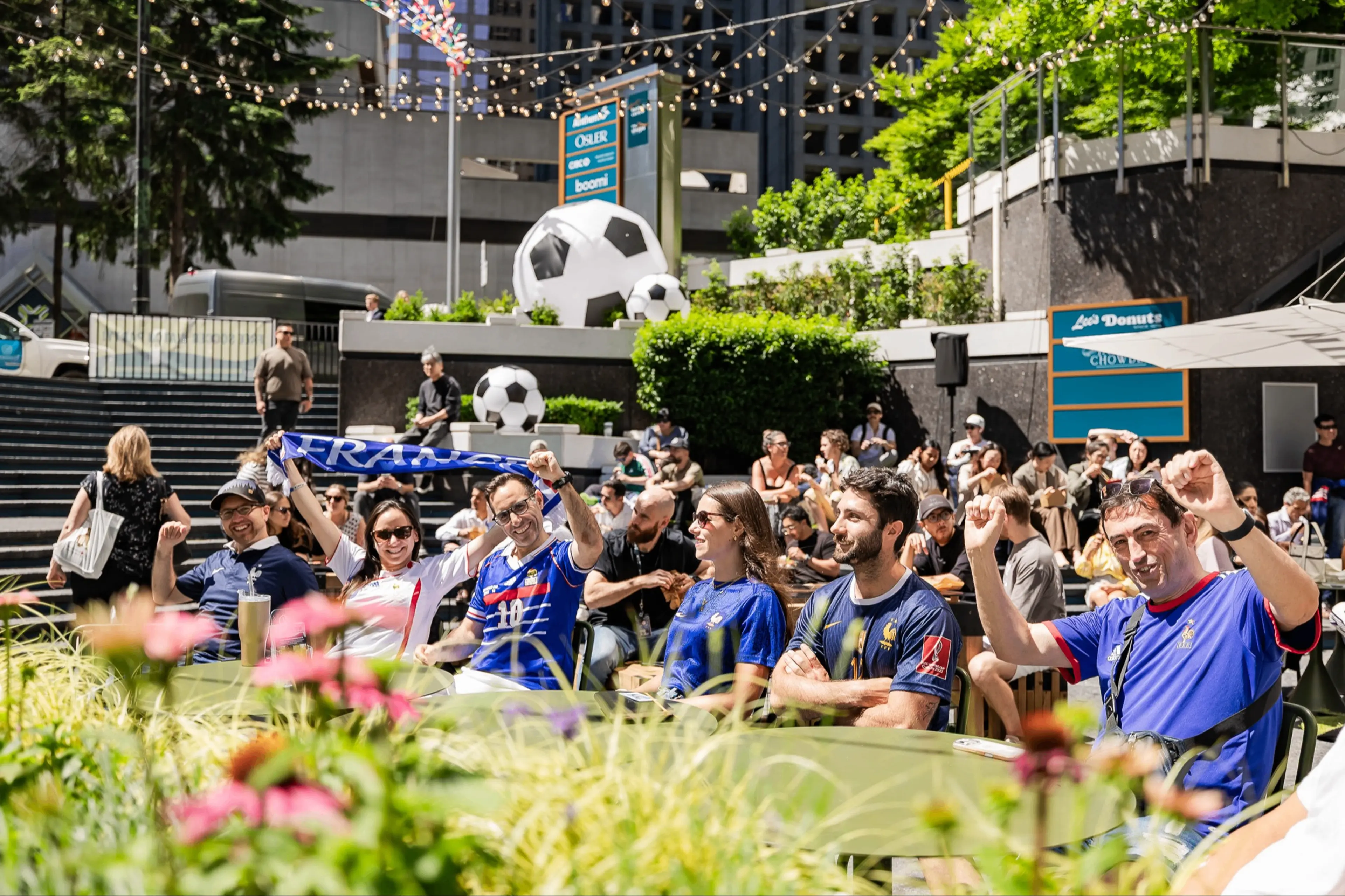 Group of people outdoors wearing soccer jerseys and cheering, with large soccer ball decorations and greenery in the background.