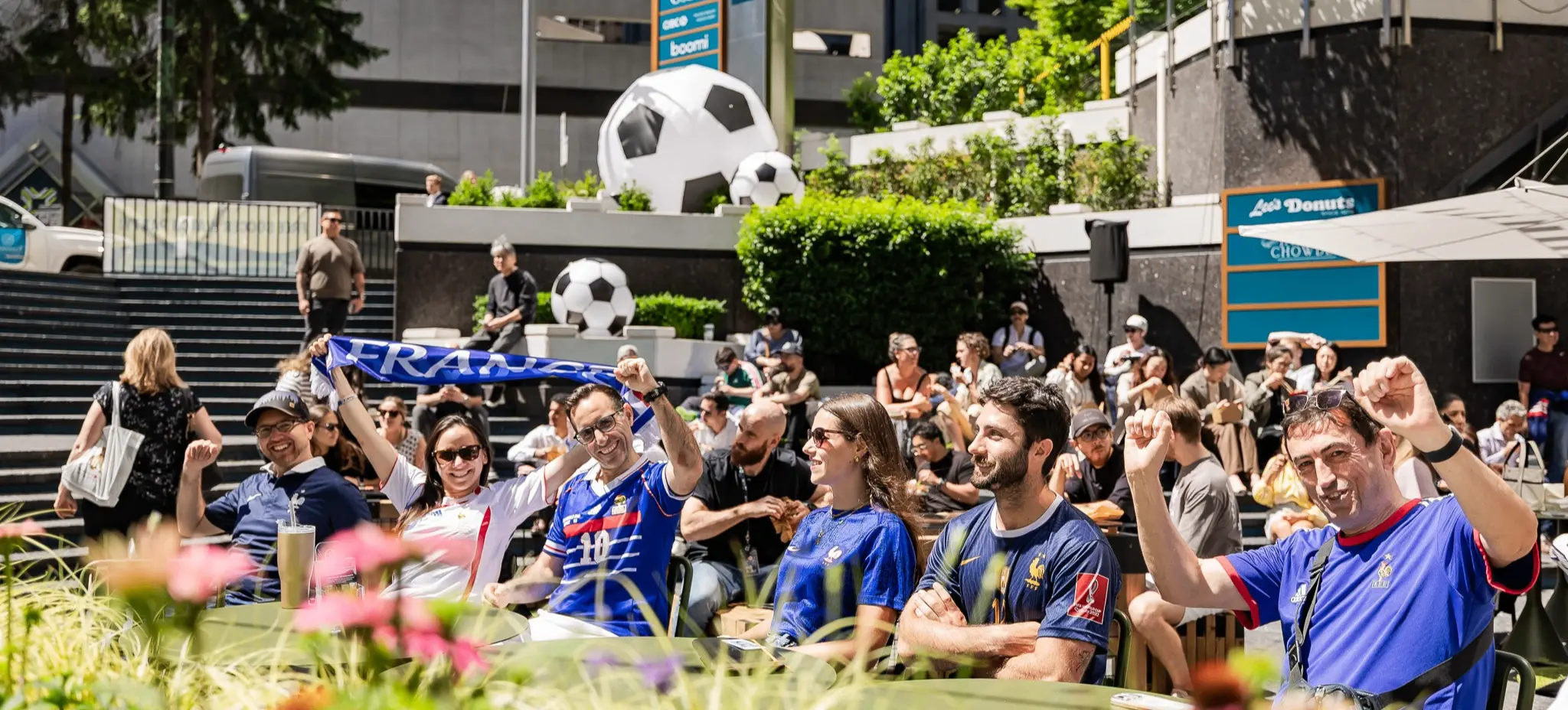 Group of people outdoors wearing soccer jerseys and cheering, with large soccer ball decorations and greenery in the background.