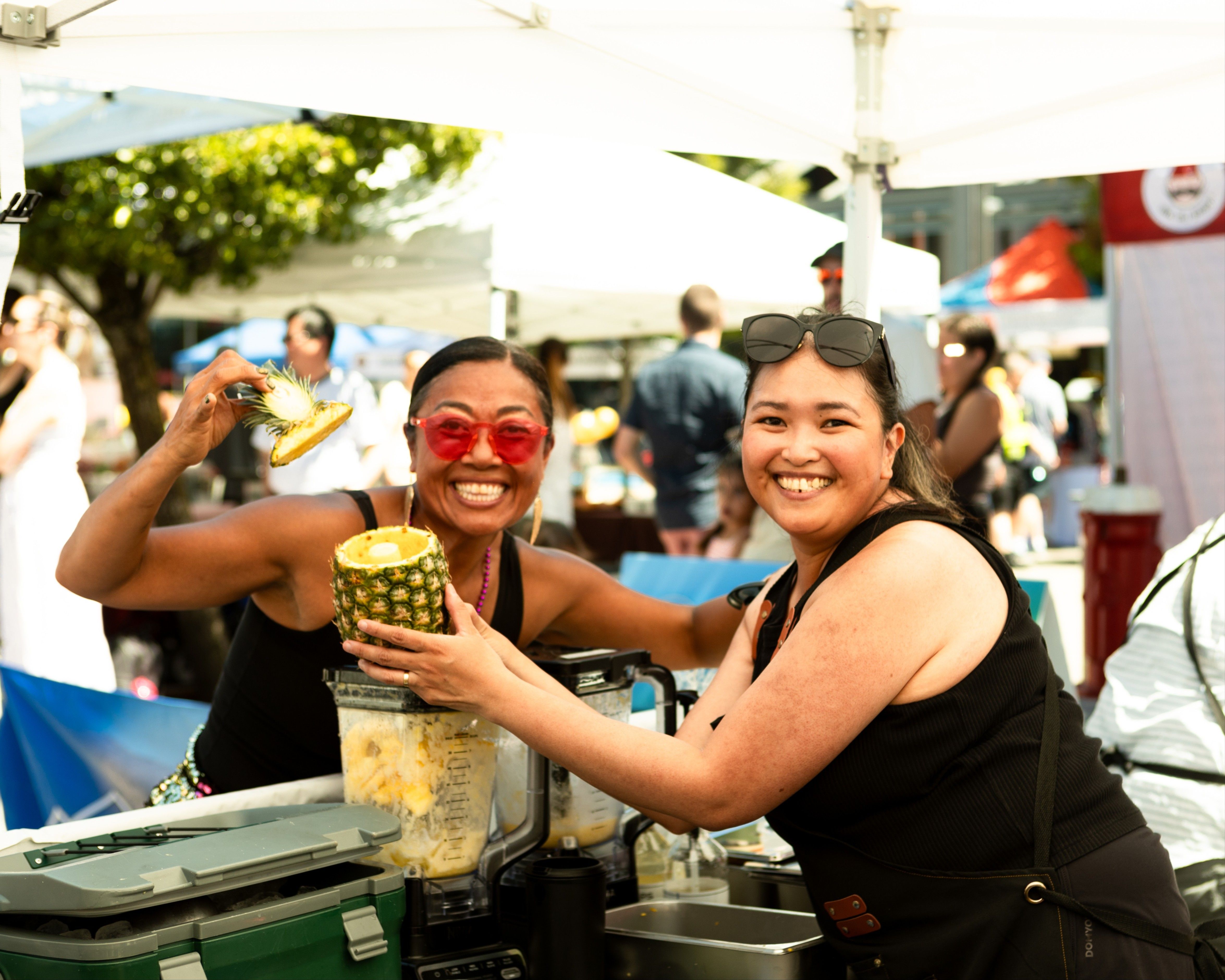 Two smiling people at an outdoor market holding a hollowed pineapple and pineapple top near a blender filled with pineapple chunks.