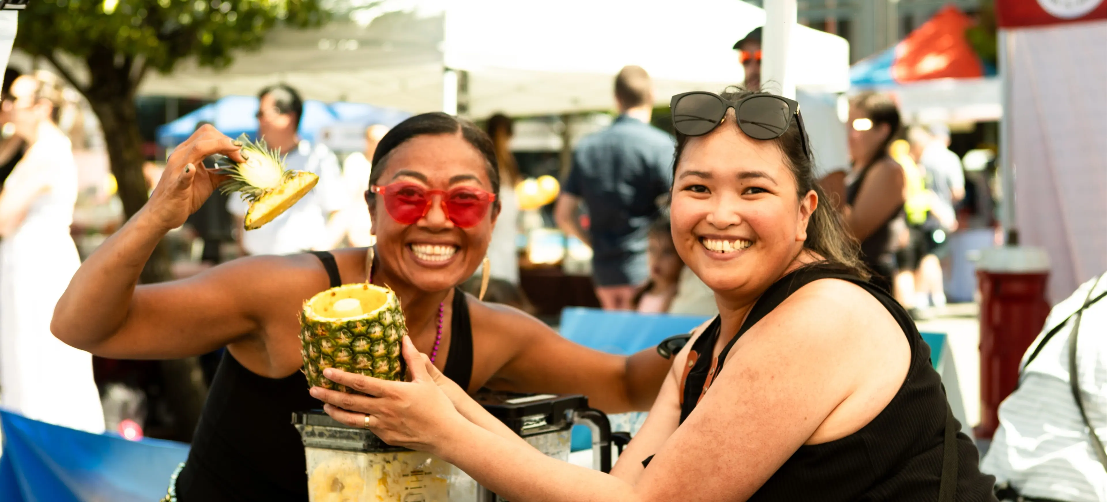 Two smiling people at an outdoor market holding a hollowed pineapple and pineapple top near a blender filled with pineapple chunks.