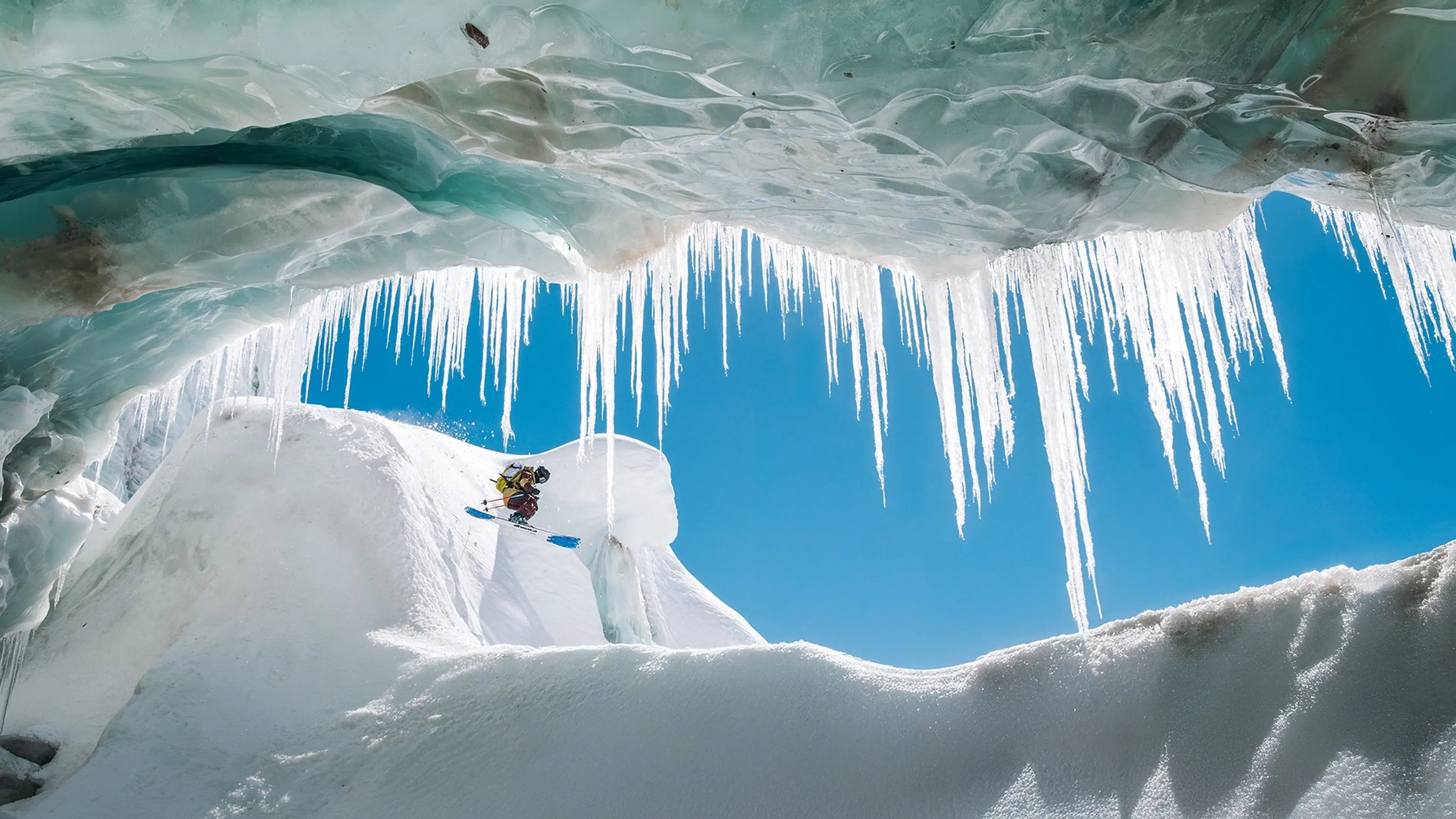 Person skiing down a snowy slope framed by large icicles hanging from an ice cave ceiling against a clear blue sky.