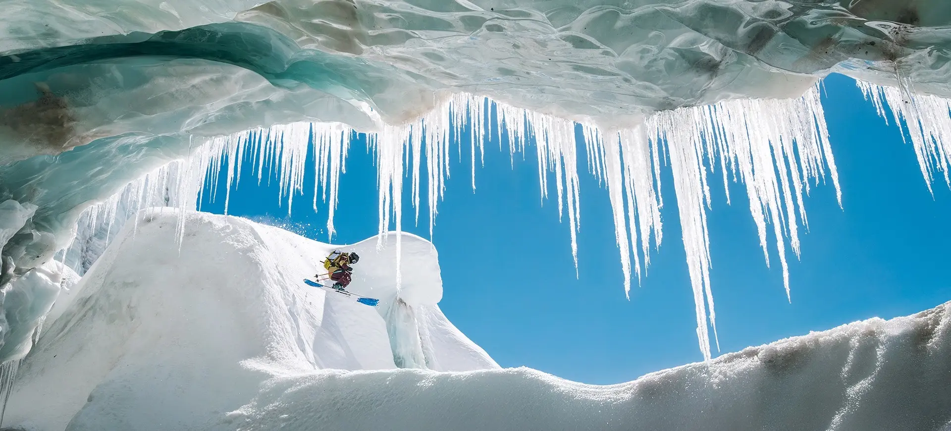 Person skiing down a snowy slope framed by large icicles hanging from an ice cave ceiling against a clear blue sky.