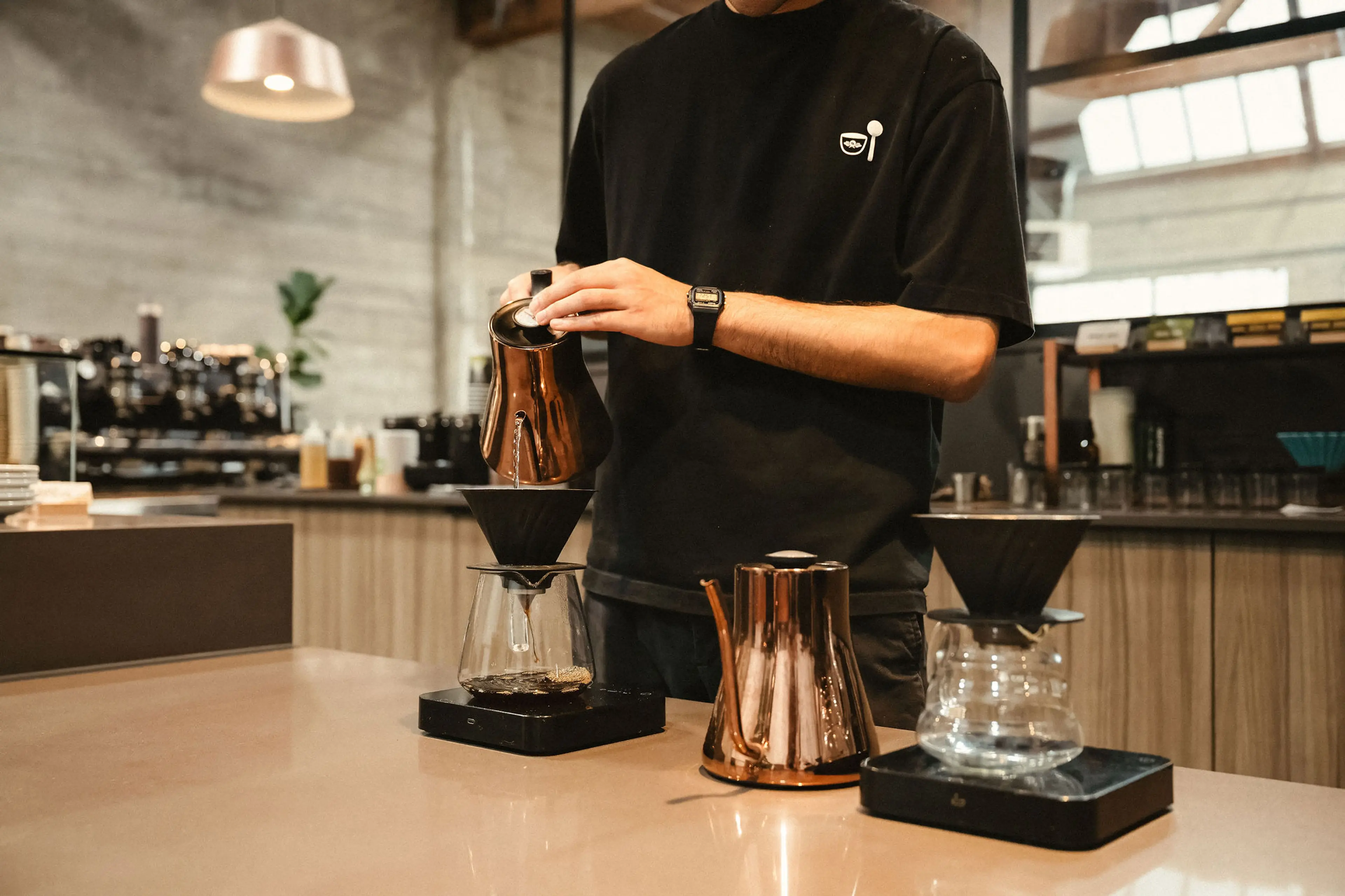 Person pouring hot water from a copper kettle into a coffee dripper on a glass carafe in a modern coffee shop.