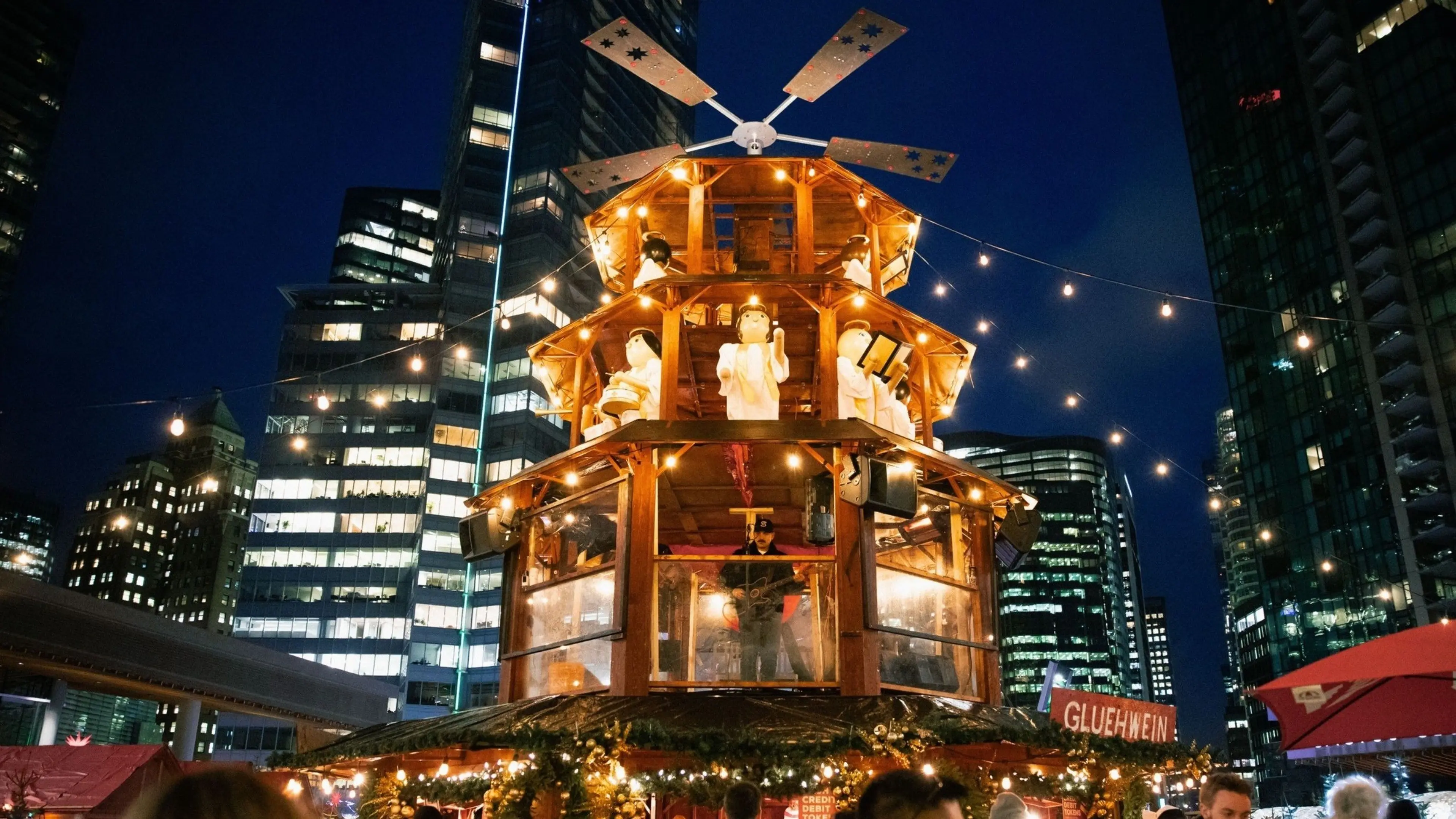 Wooden Christmas pyramid decorated with lights and angel figurines, set against a city skyline at night with people nearby.