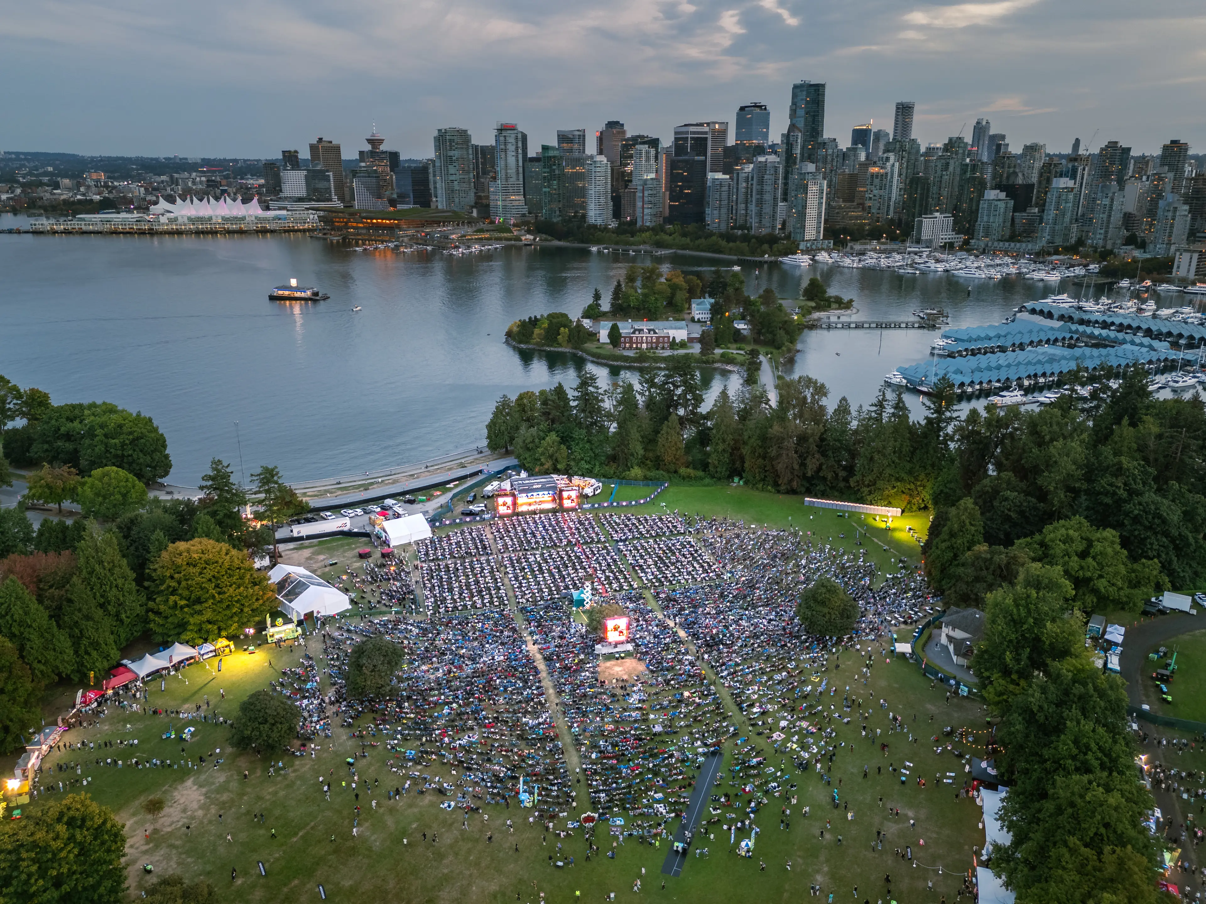 A large outdoor concert with a crowd seated on grass near water, with a city skyline in the background at dusk.