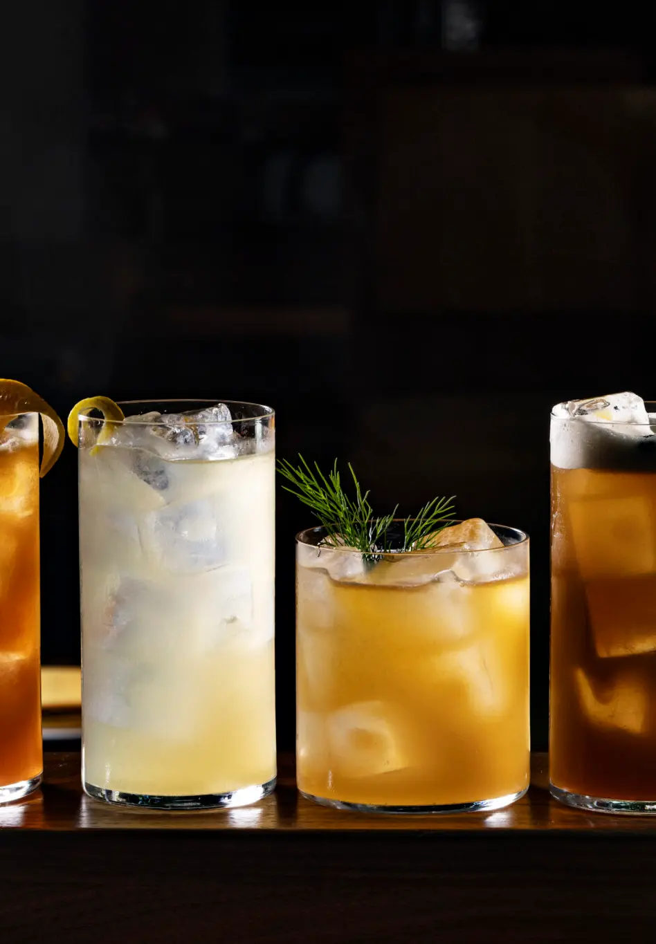 Four iced cocktails in different glasses on a wooden surface with dark background and some green leaves on the right.