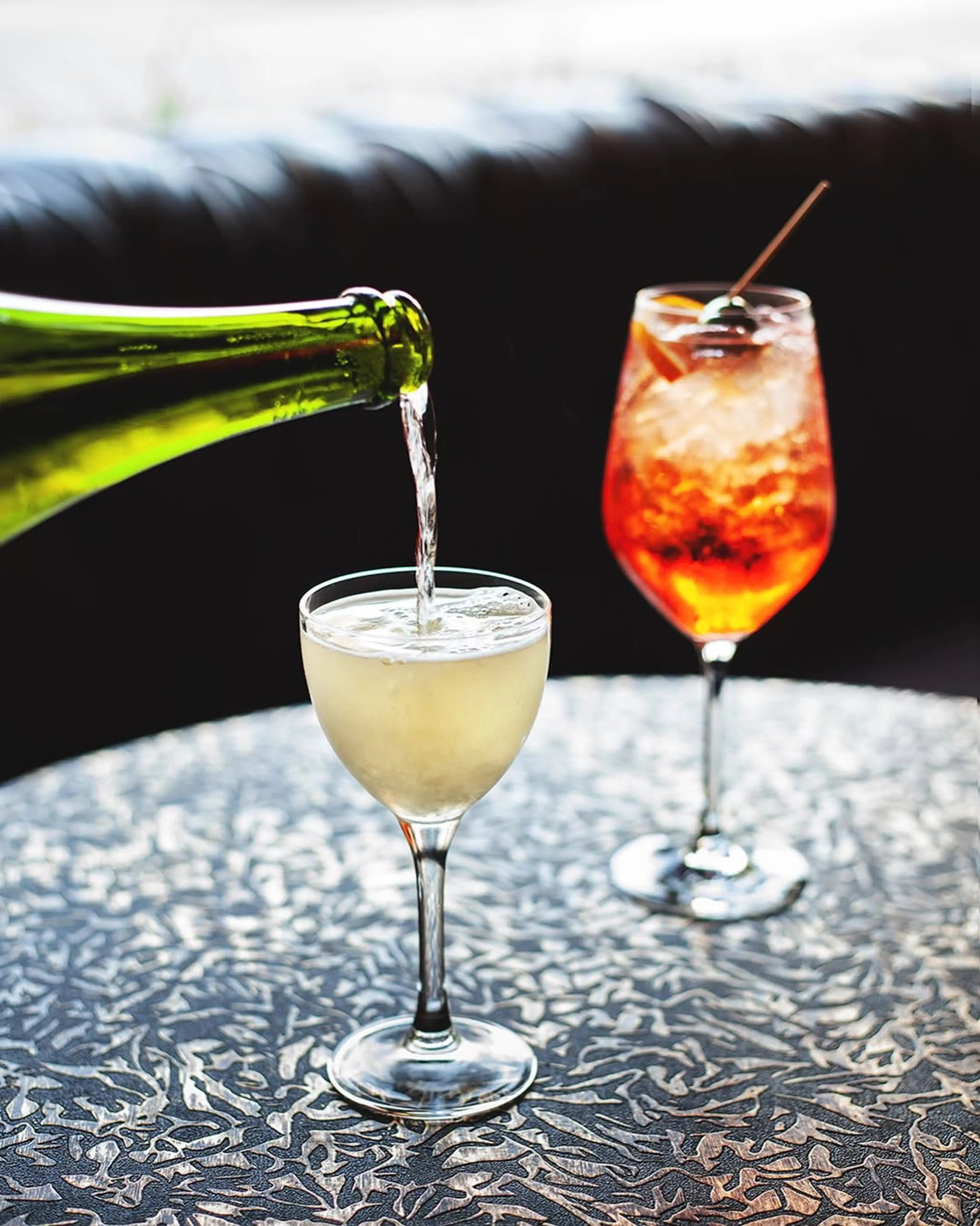 A green bottle pouring a clear drink into a stemmed glass on a patterned table, with an orange cocktail garnished with a cherry in the background.