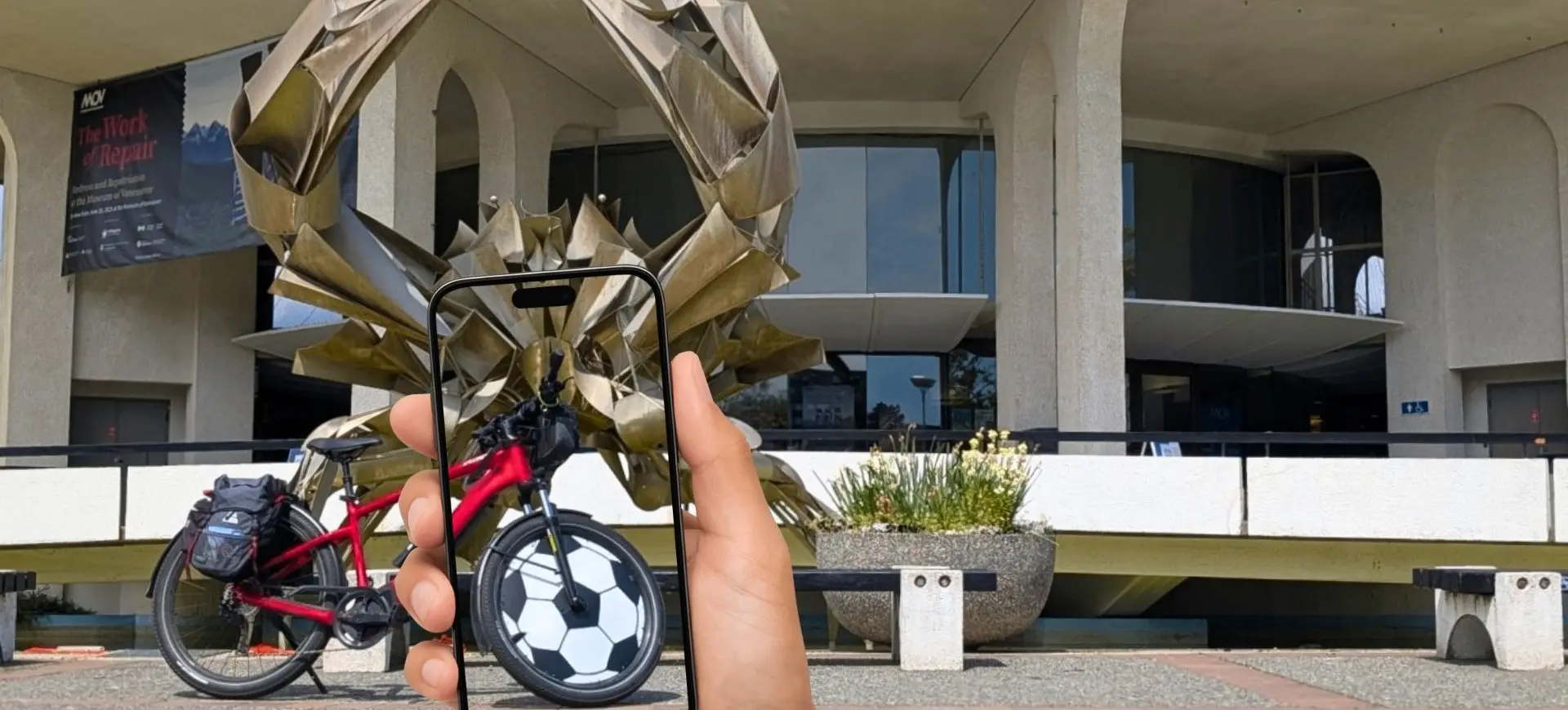 Hand holding a phone showing a soccer ball image aligned with the front wheel of a red bicycle in front of a large crab sculpture and a building.