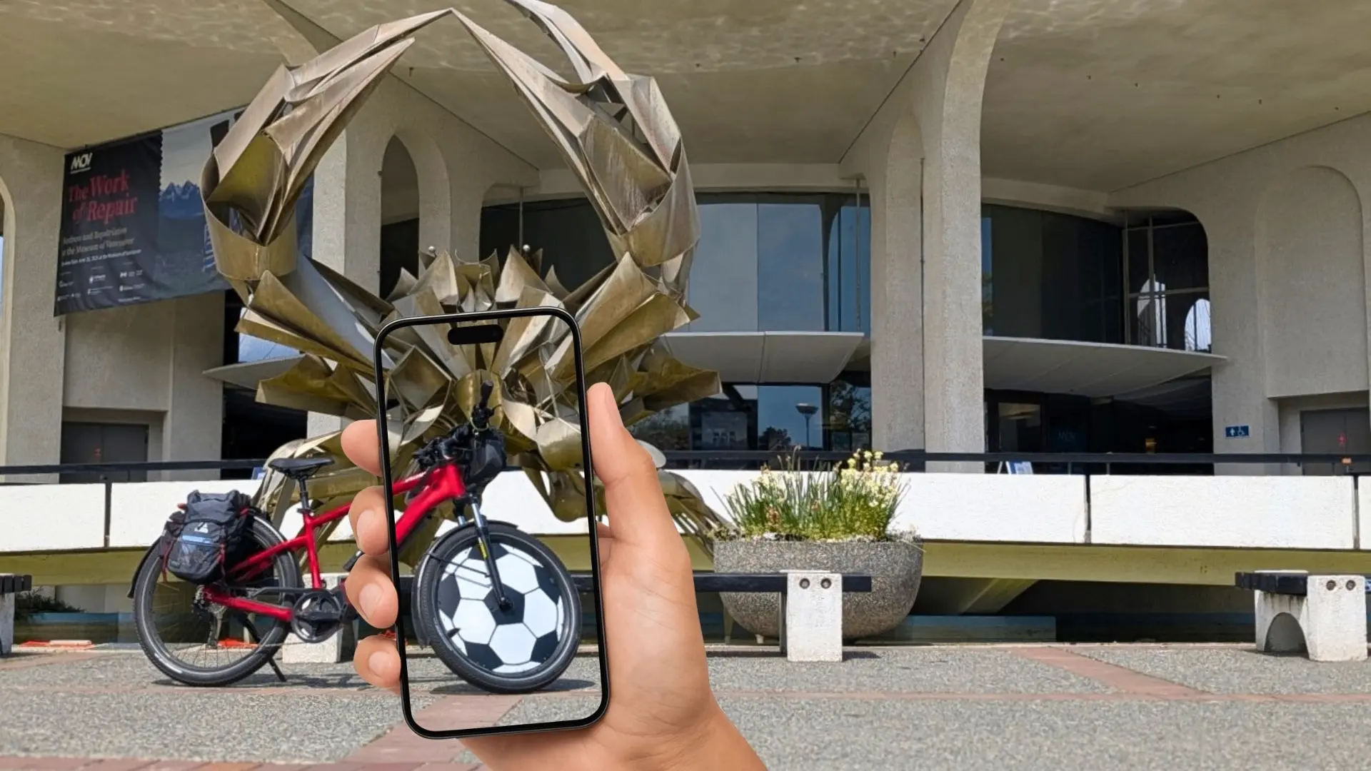 Hand holding a phone showing a soccer ball image aligned with the front wheel of a red bicycle in front of a large crab sculpture and a building.