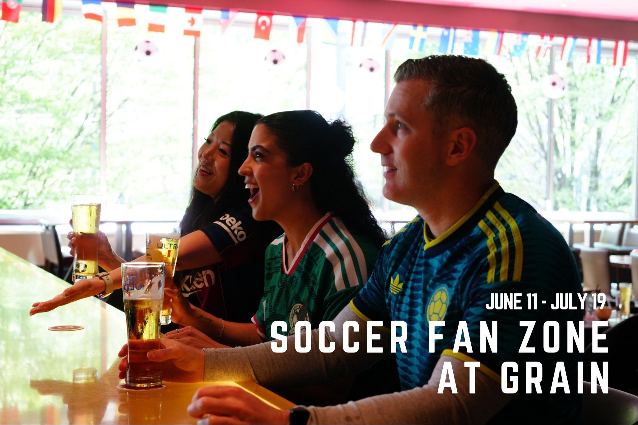 Three people in soccer jerseys sitting at a bar holding glasses of beer with international flags hanging above, text reads June 11 - July 19 Soccer Fan Zone at Grain.