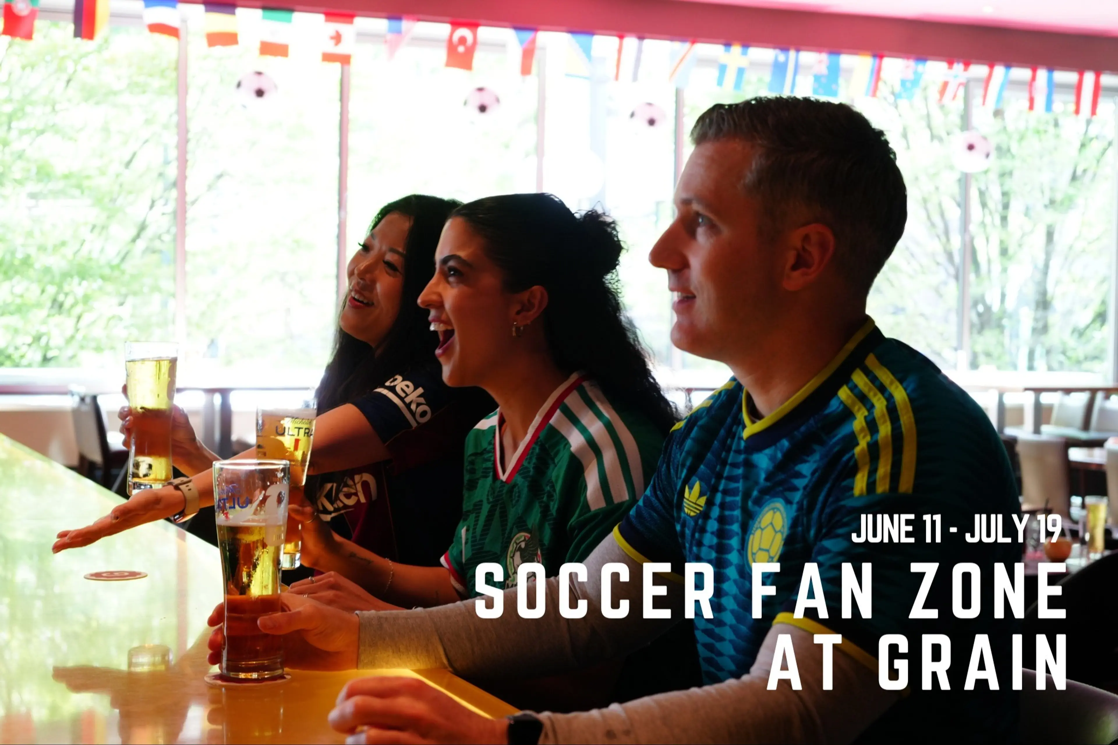 Three people in soccer jerseys sitting at a bar holding glasses of beer with international flags hanging above, text reads June 11 - July 19 Soccer Fan Zone at Grain.