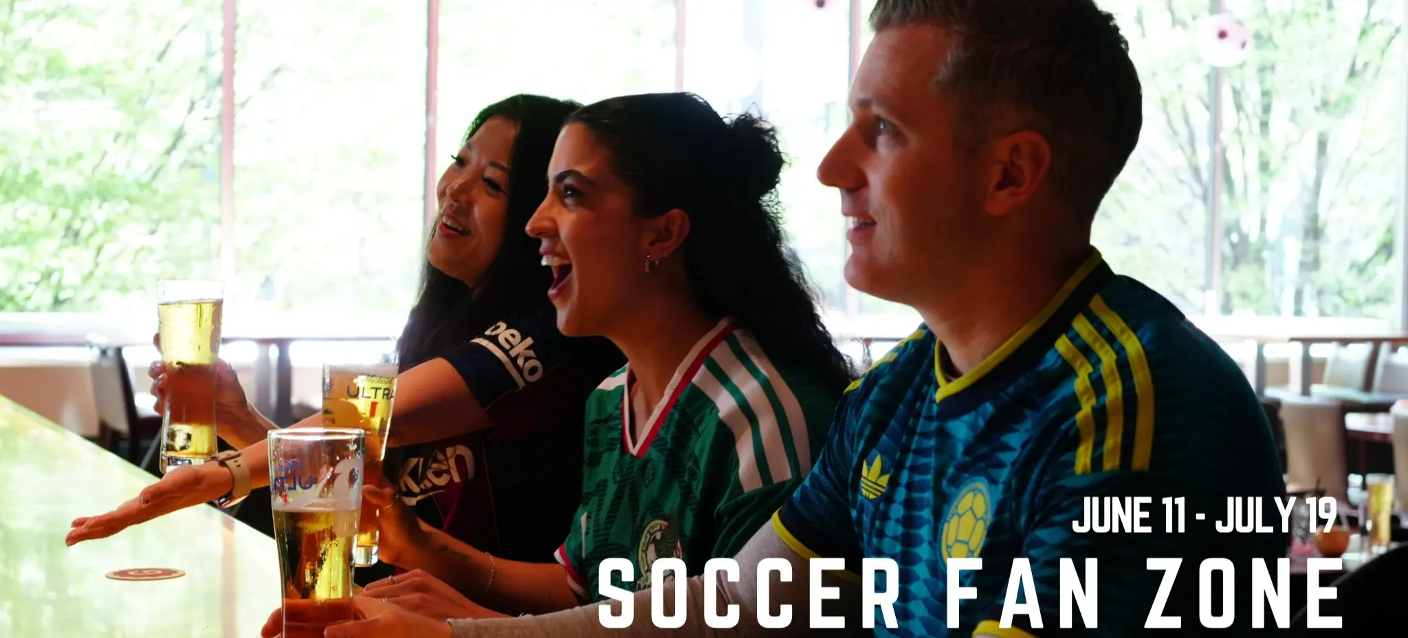 Three people in soccer jerseys sitting at a bar holding glasses of beer with international flags hanging above, text reads June 11 - July 19 Soccer Fan Zone at Grain.