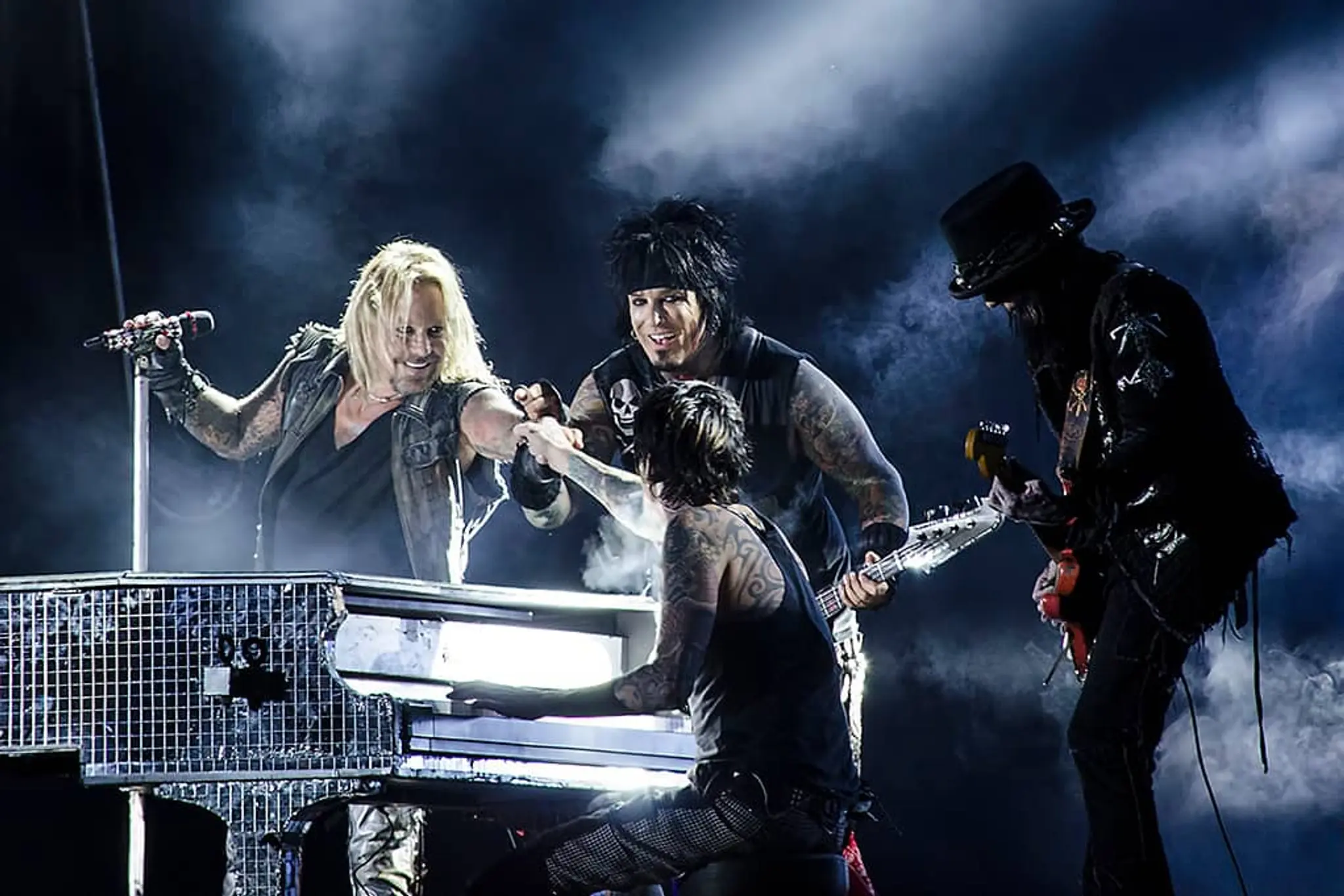 Four rock musicians performing on stage with smoke effects, one playing piano, one holding microphone, and two playing guitars.