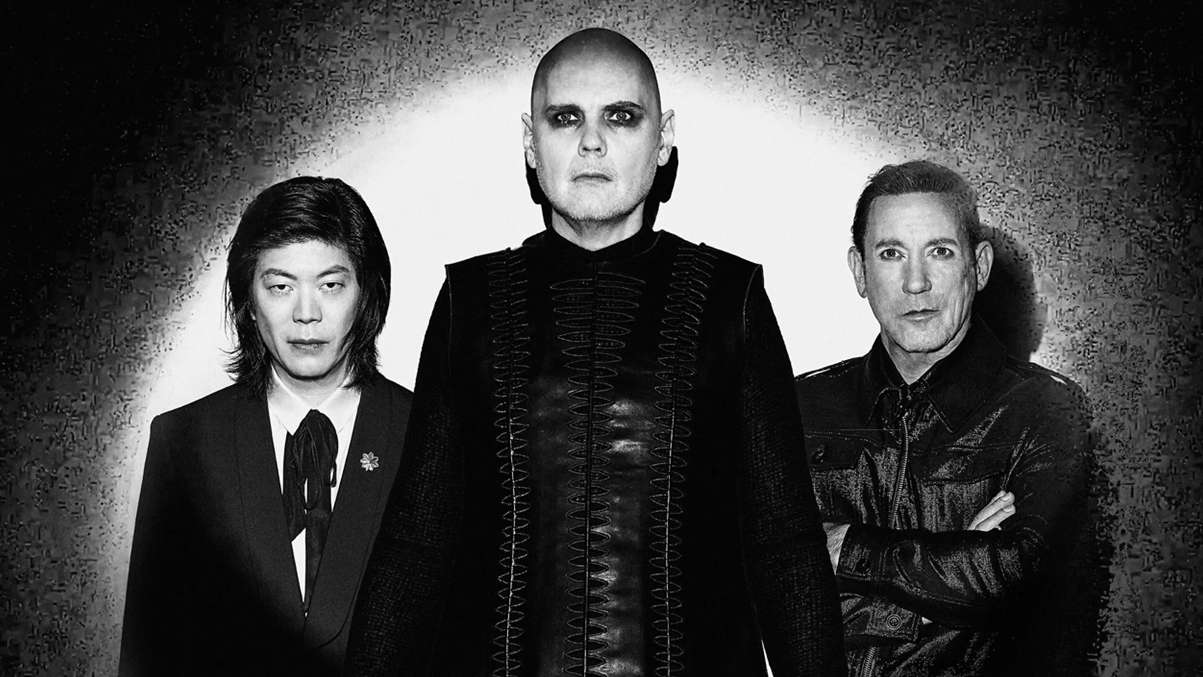 Three people in dark clothing standing against a textured wall with spotlight behind them, one with bald head and dark eye makeup.