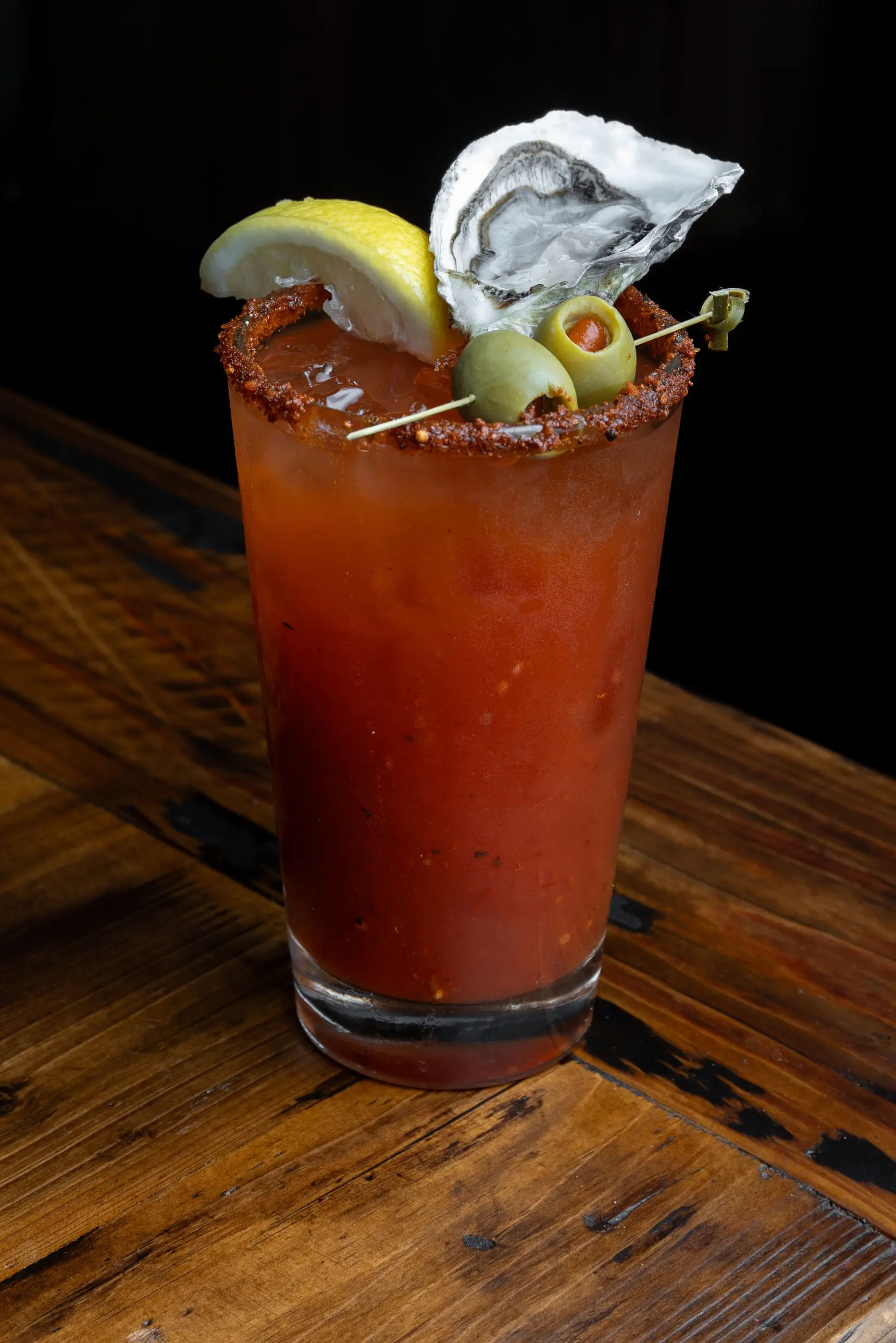 Bloody Mary cocktail garnished with lemon wedge, two green olives, and an oyster on a wooden table.