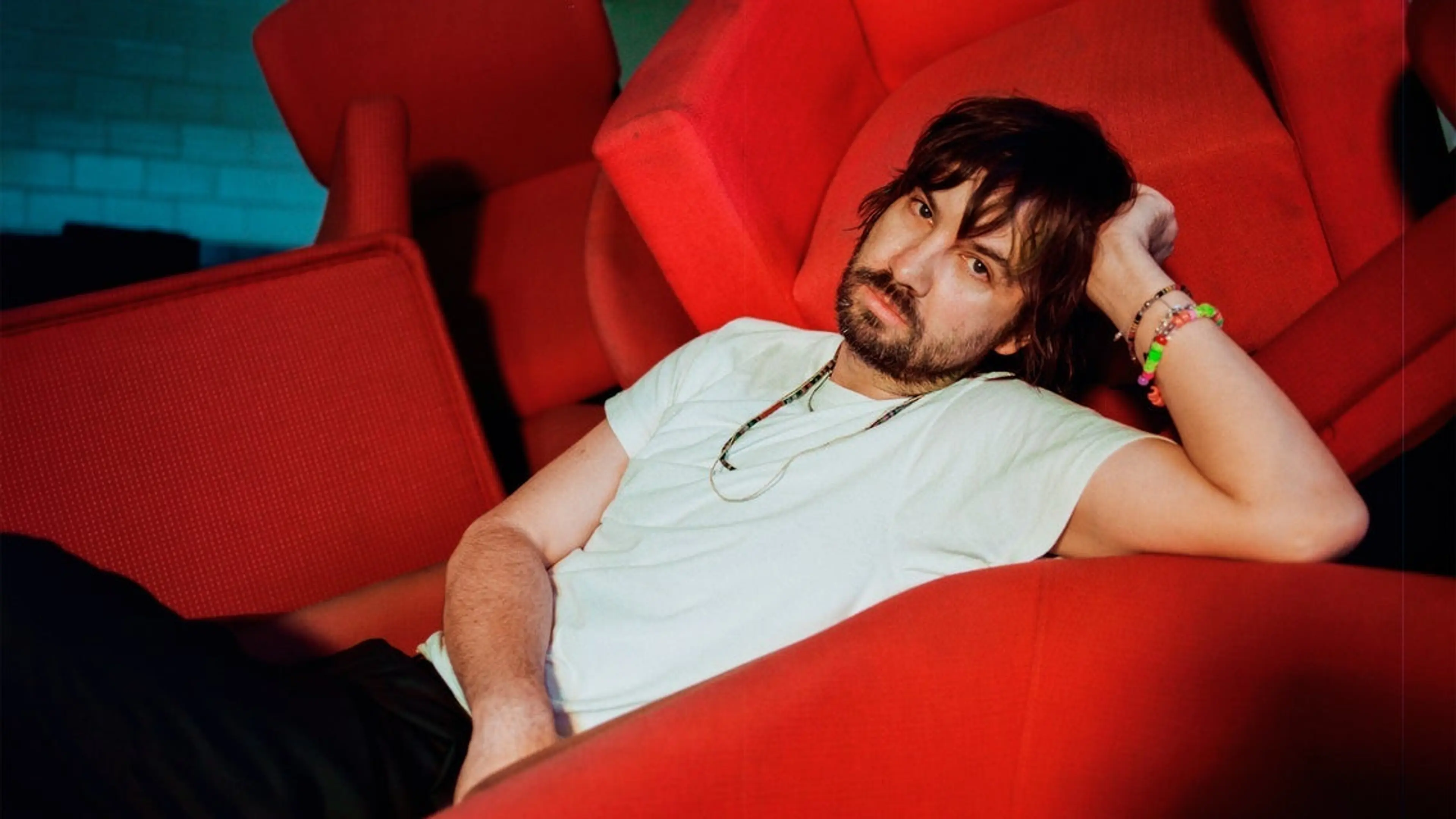 Person with dark hair and beard reclining on a red chair, wearing a white t-shirt and colorful bracelets.