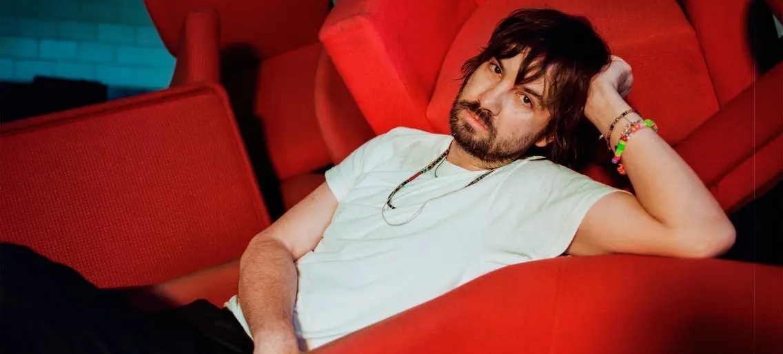 Person with dark hair and beard reclining on a red chair, wearing a white t-shirt and colorful bracelets.