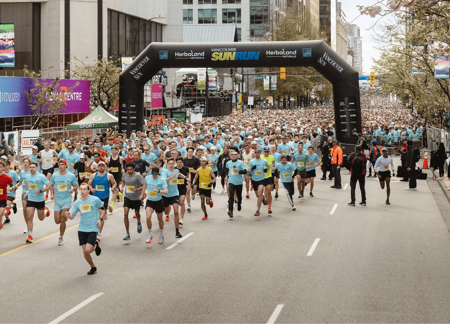Large group of runners starting the Vancouver Sun Run under a black inflatable arch on a city street.