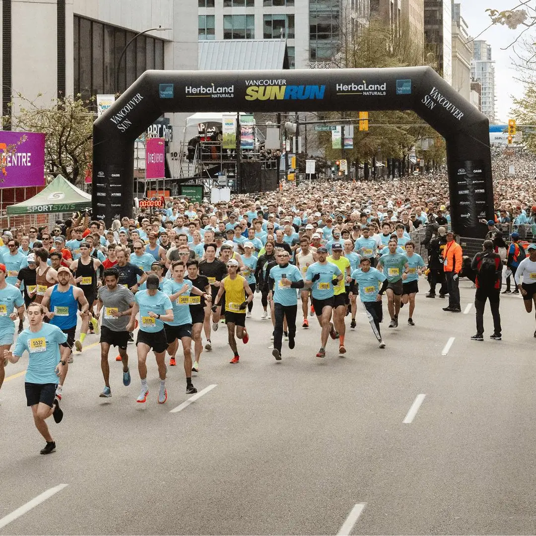 Large group of runners starting the Vancouver Sun Run under a black inflatable arch on a city street.