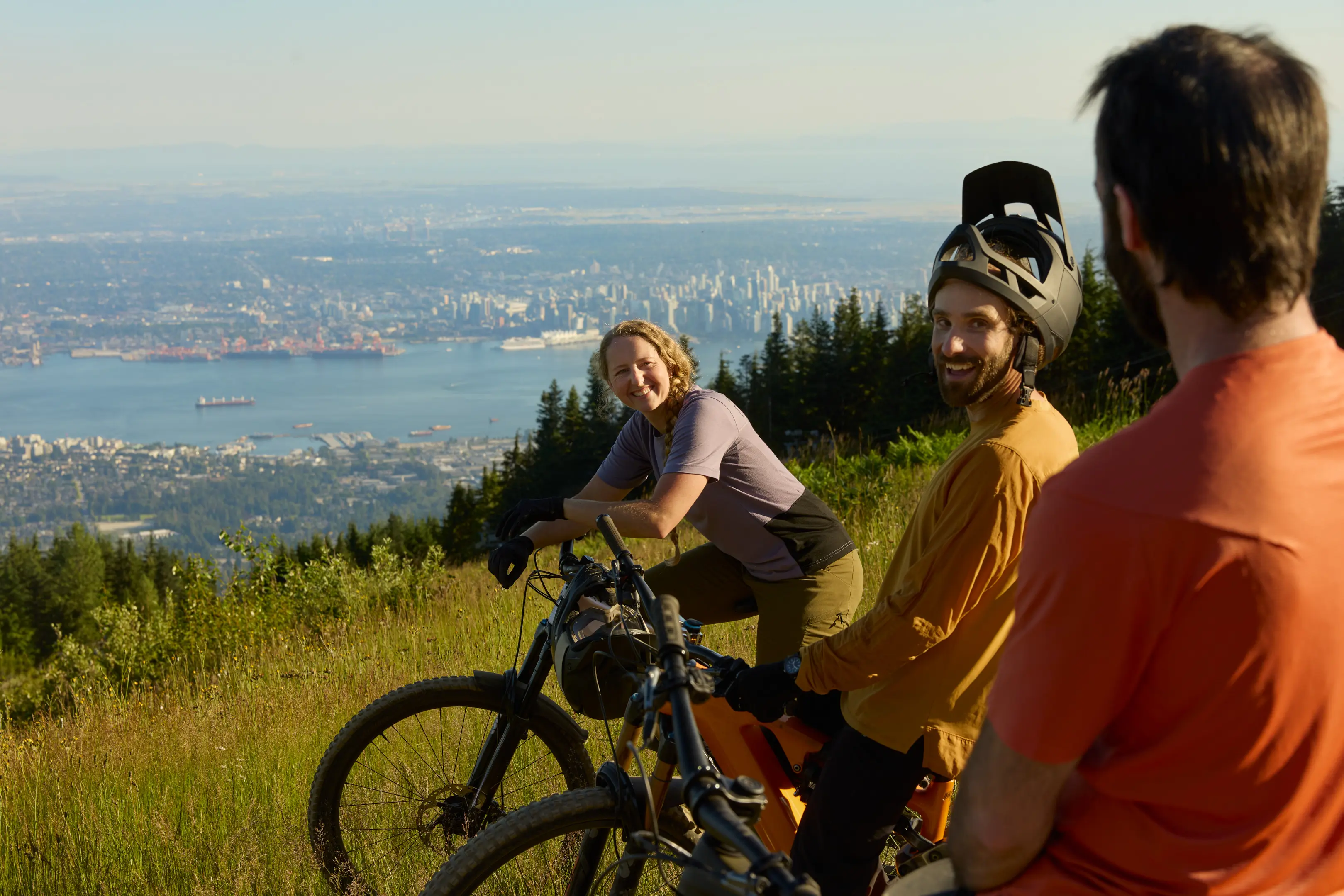 Three people with mountain bikes on a grassy hill overlooking a city and water, two smiling and one facing away.