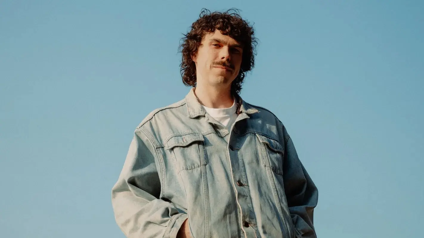 Person with curly hair and mustache wearing a light denim jacket against a clear blue sky.