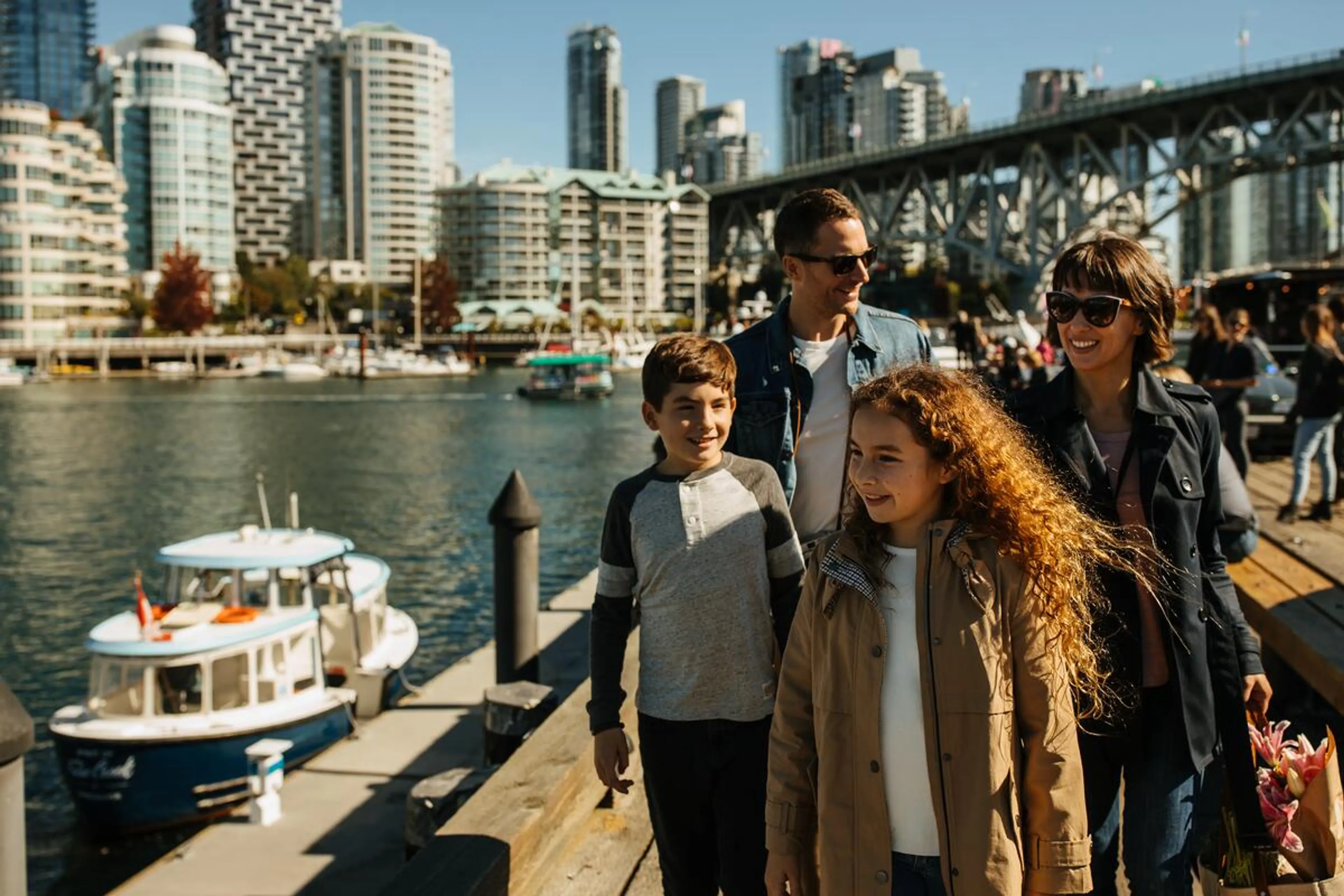 A group of four people walking on a waterfront dock with boats and city buildings in the background on a sunny day.