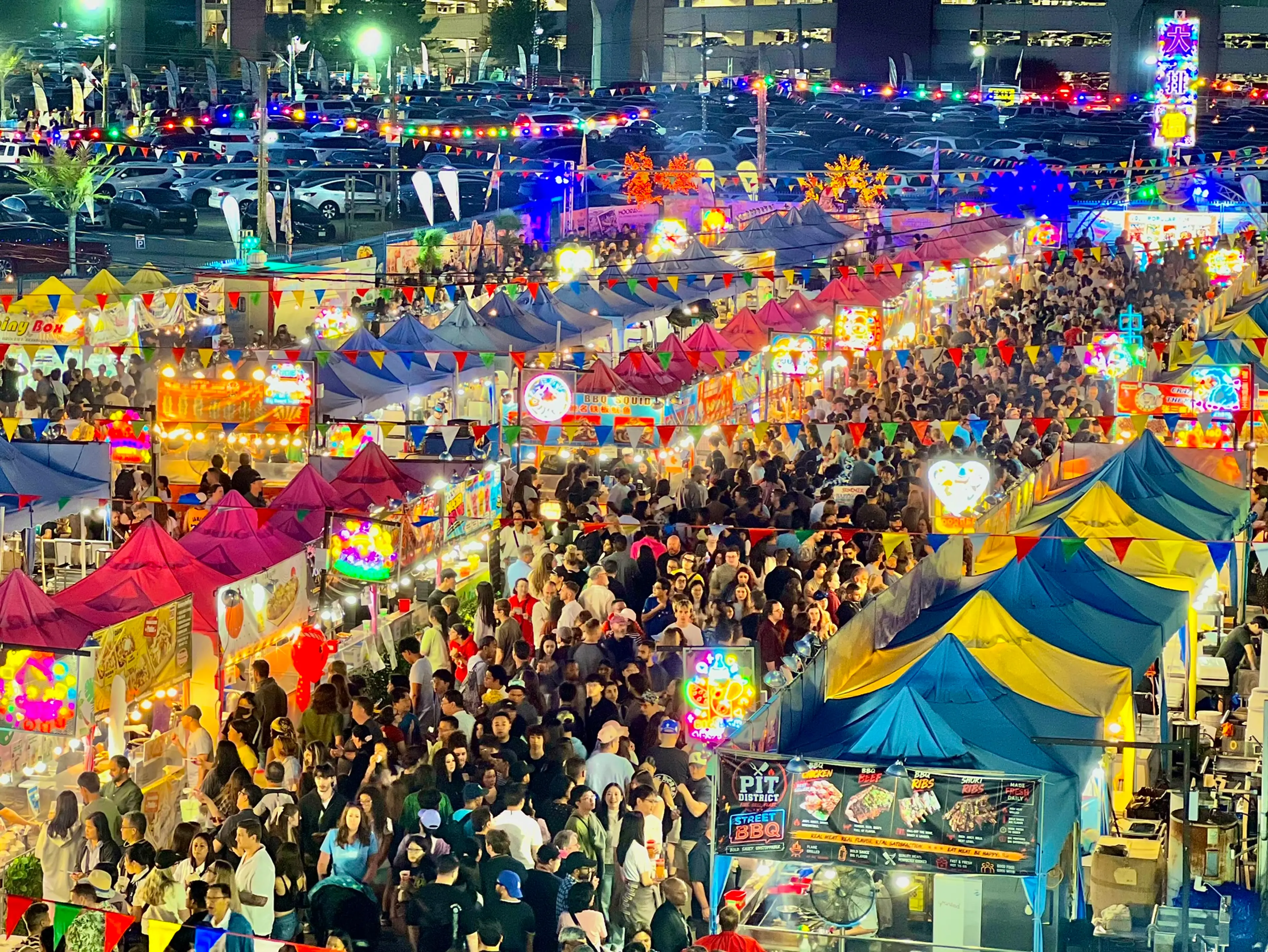 Crowded night market with colorful tents, bright lights, and many people walking between stalls.