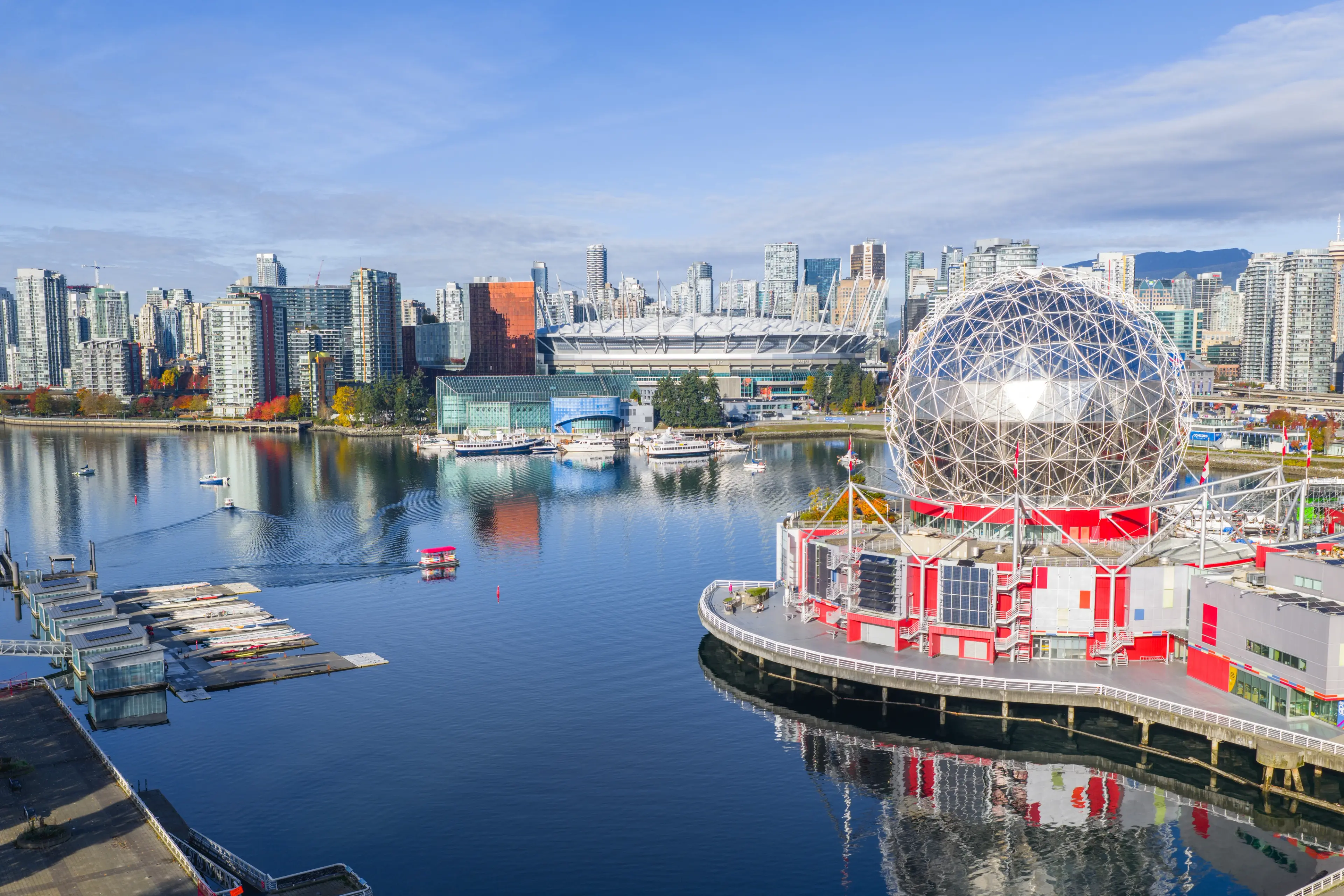 Vancouver city skyline with modern buildings, the large geodesic dome of Science World, and boats on calm water under a blue sky.