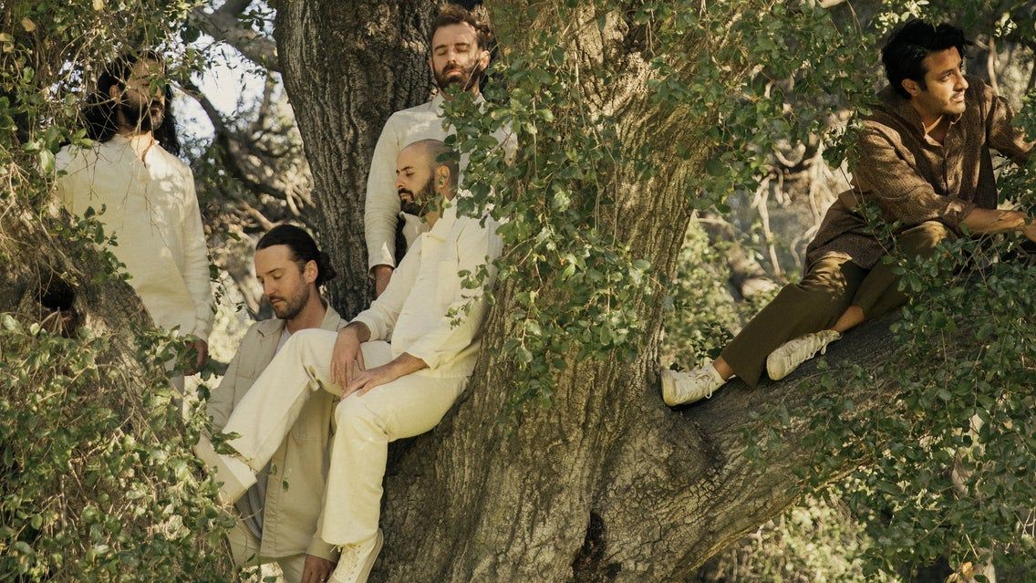 Five people sitting and standing on a large tree with green leaves, dressed in light and earth-toned clothing.