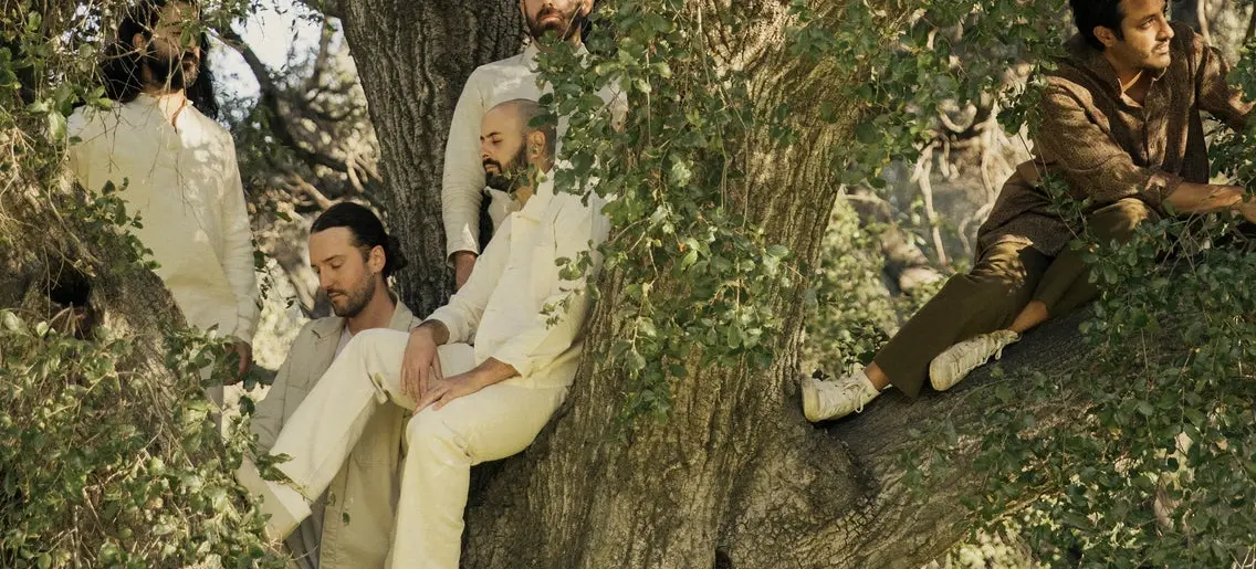 Five people sitting and standing on a large tree with green leaves, dressed in light and earth-toned clothing.