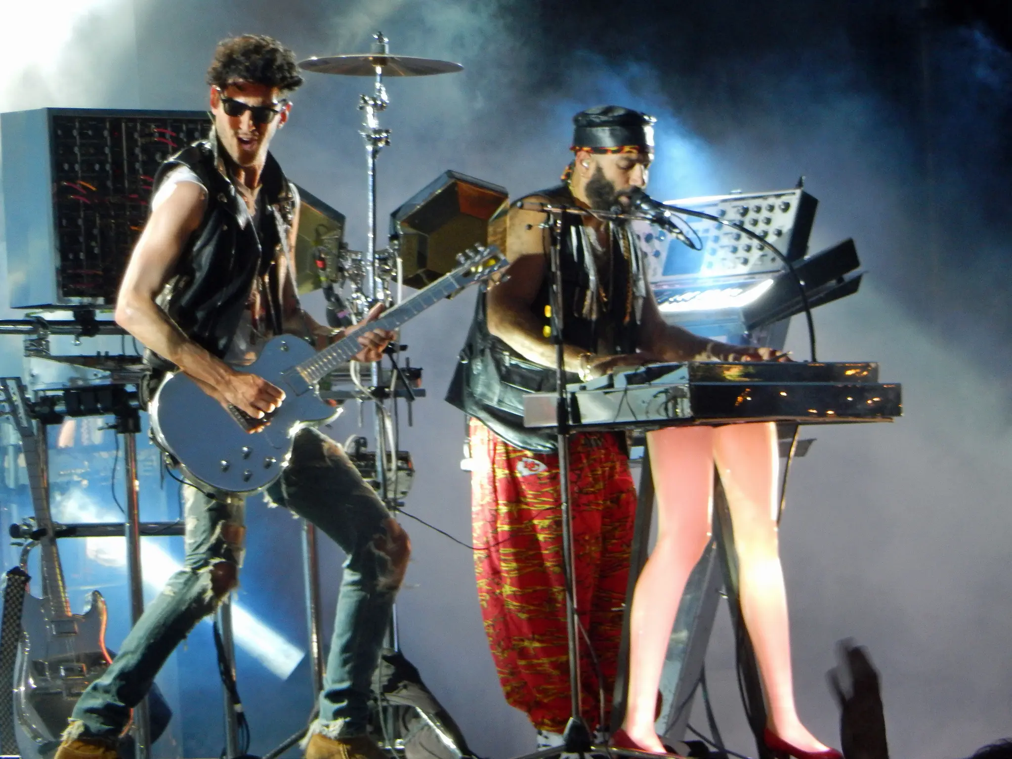 Two male musicians performing on stage, one playing electric guitar and the other playing keyboard with microphone, stage lights and smoke in background.