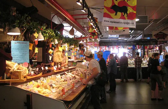 People shopping at a market stall with various cheeses and sausages displayed under hanging lights and greenery.