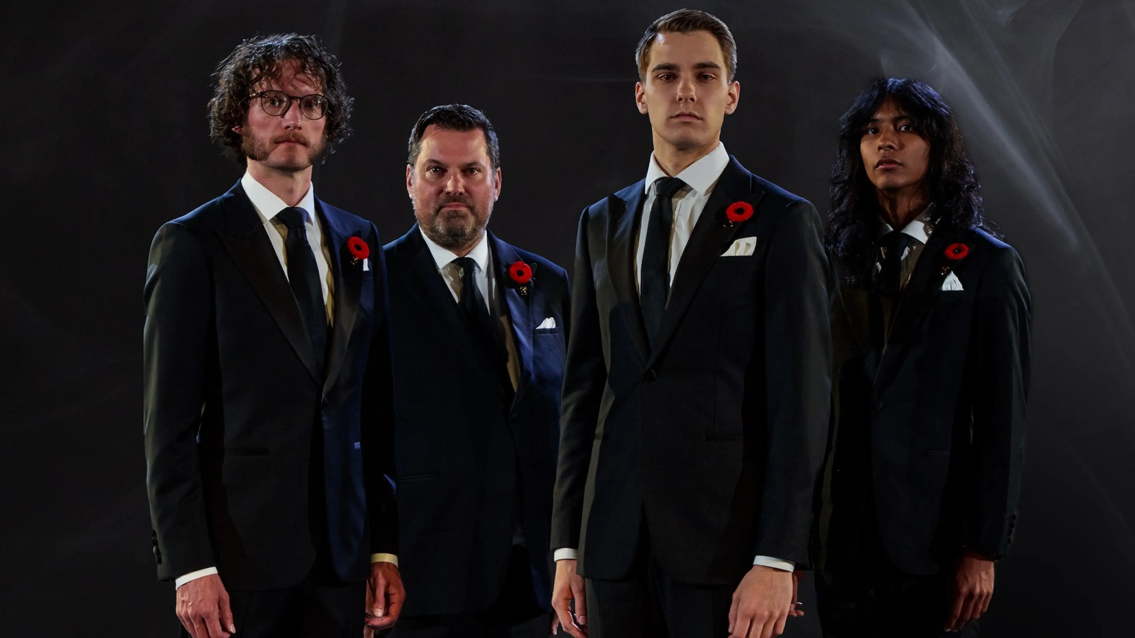 Four people in black suits with white shirts and black ties, each wearing a red poppy pin, standing against a dark background.