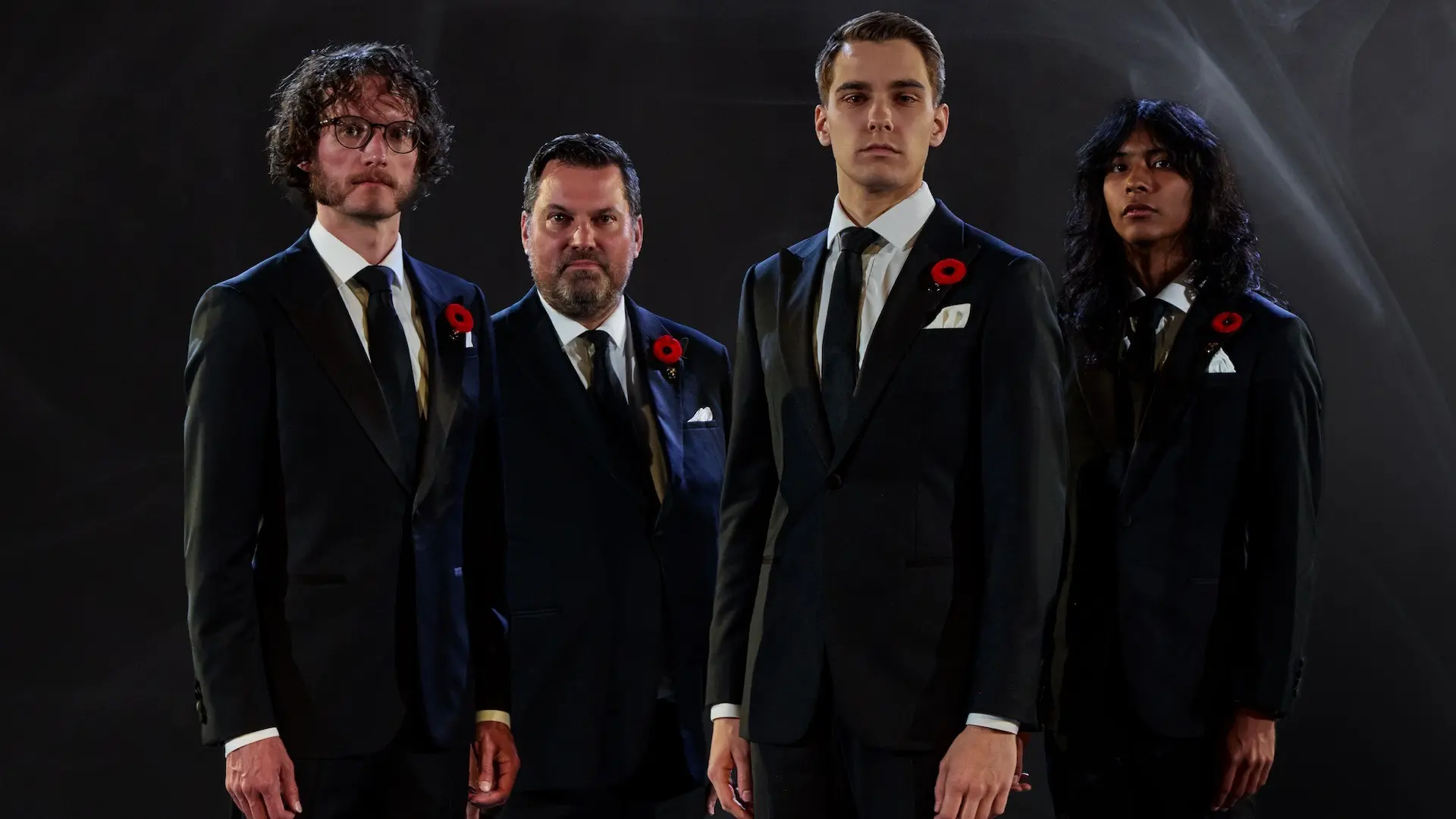 Four people in black suits with white shirts and black ties, each wearing a red poppy pin, standing against a dark background.