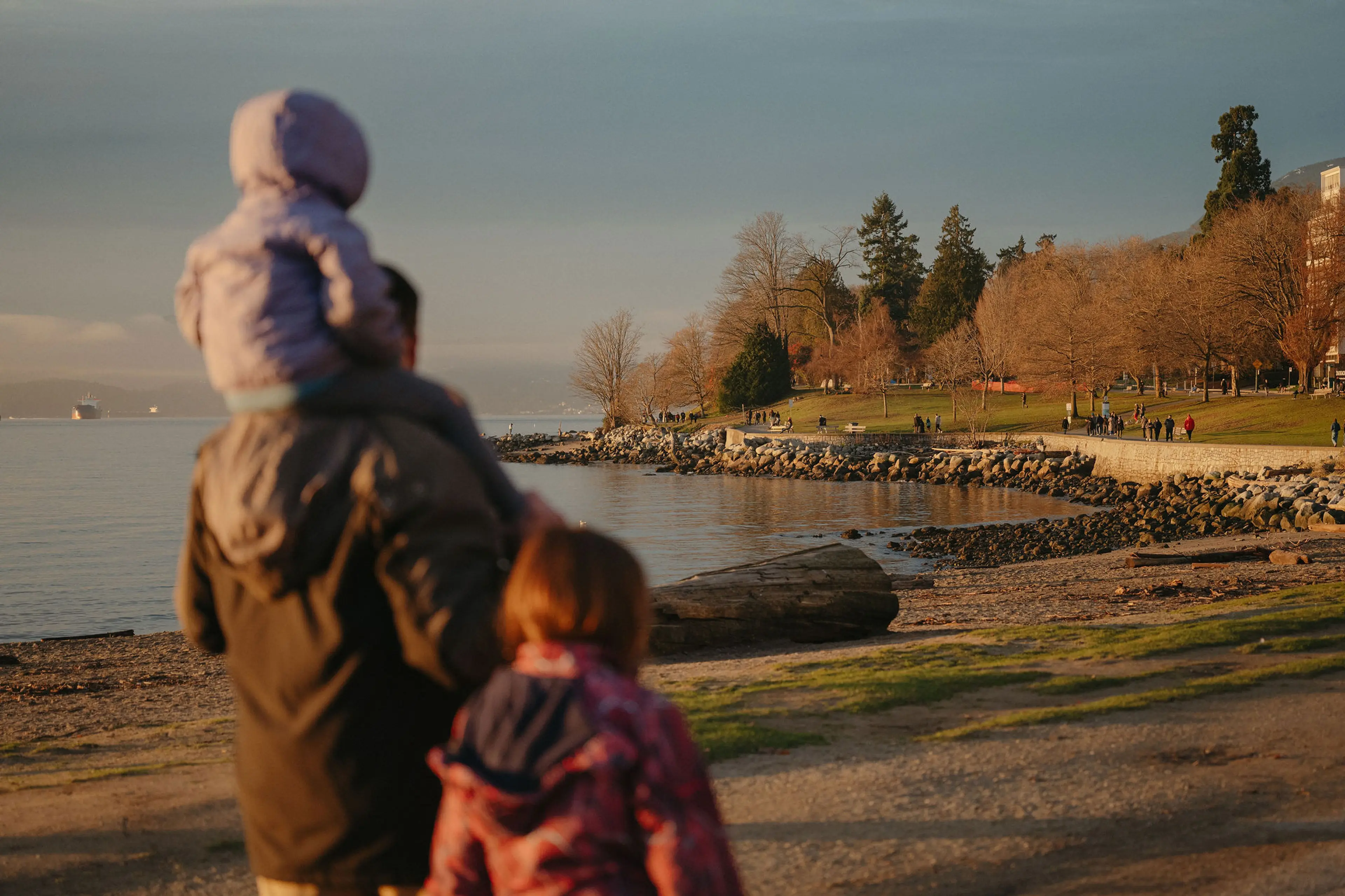 A person carrying a child on their shoulders and another child walking beside them near a rocky shoreline with a park and trees in the background.