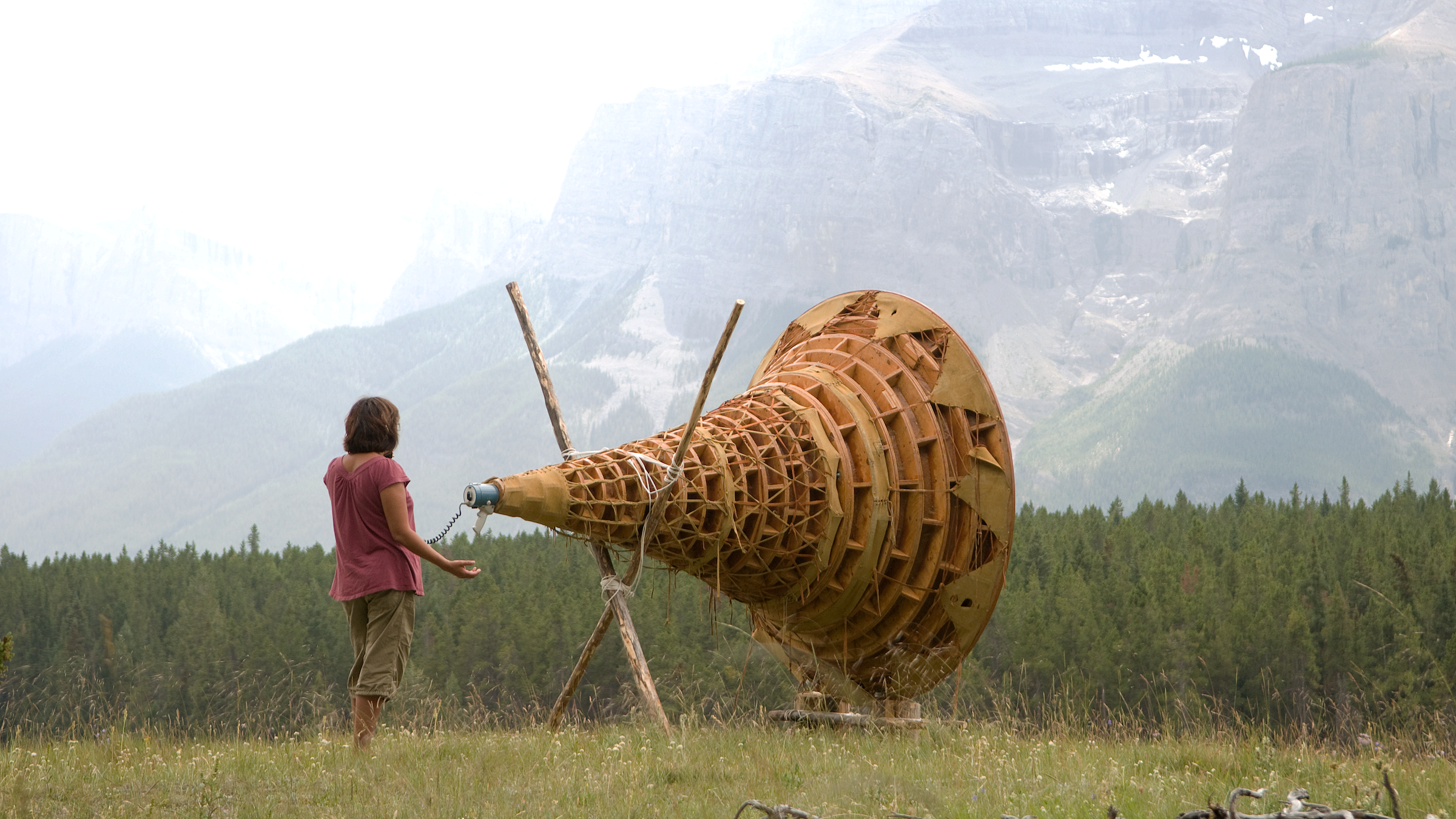 Person standing in front of trees and mountain communicating through a large wooden device
