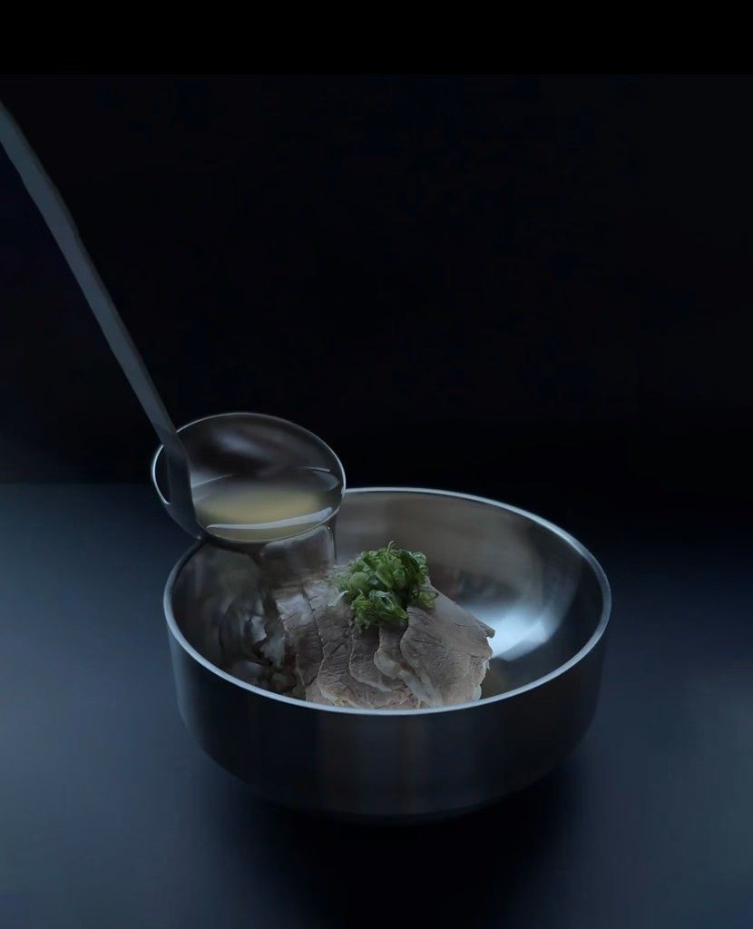 Ladle pouring broth into a metal bowl with sliced meat and chopped green onions on top.