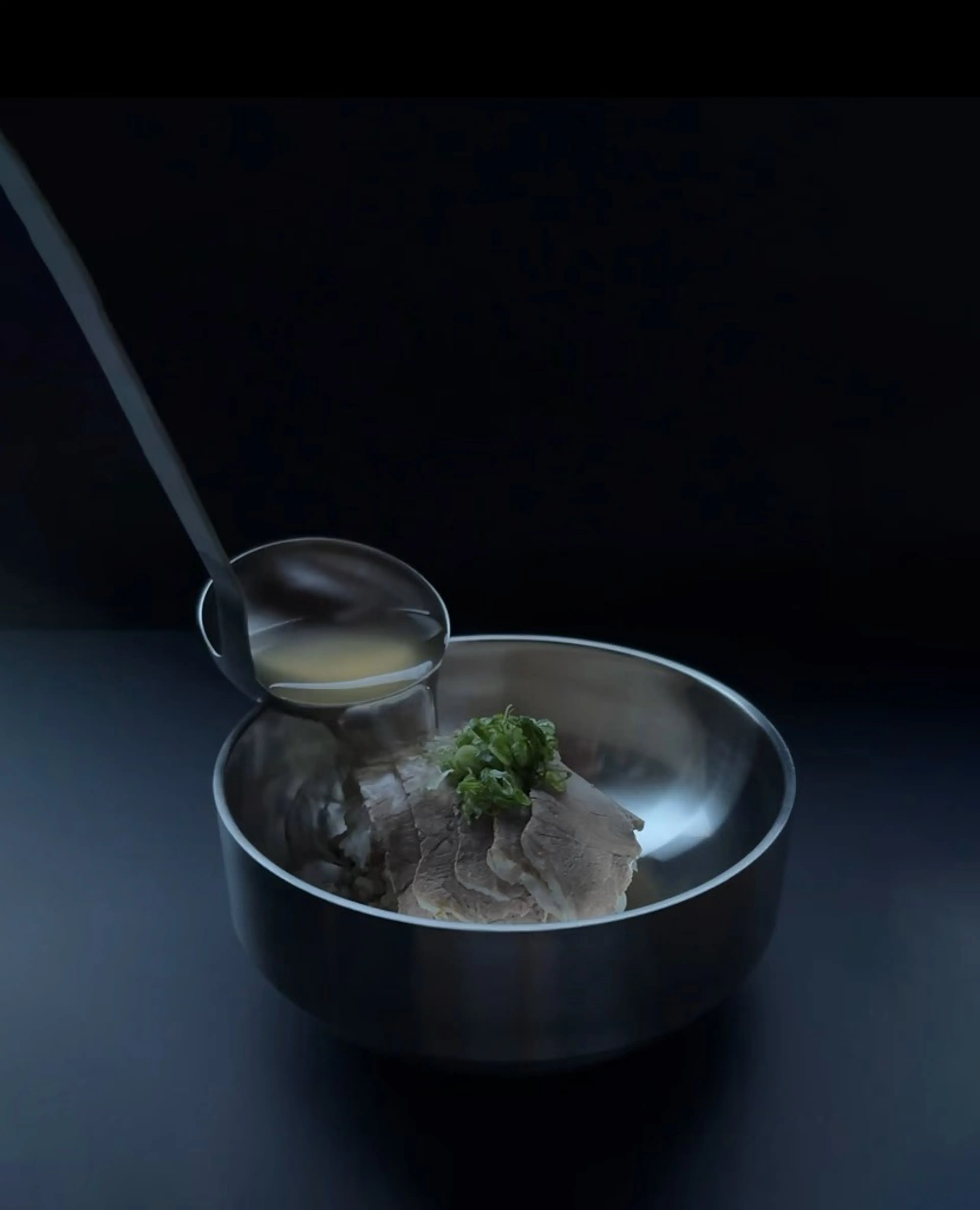 Ladle pouring broth into a metal bowl with sliced meat and chopped green onions on top.